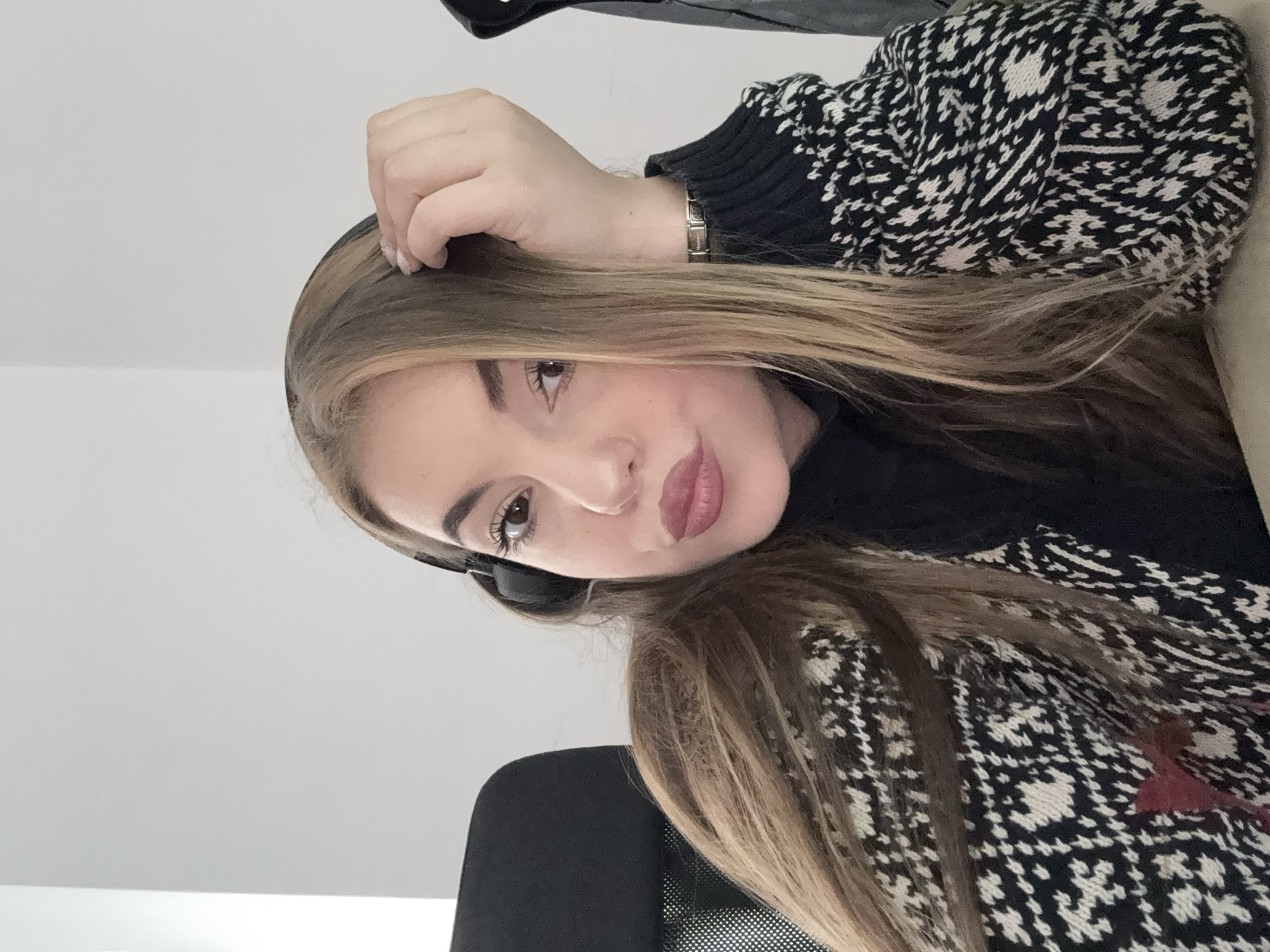 A woman with long, blonde-streaked hair is looking directly at the camera. She is wearing a black and white patterned sweater. Her right hand is partially raised to her hair. She is wearing headphones around her neck. The background is a plain white wall. A portion of a black chair is visible in the lower left. There is no evidence indicating that this image was taken in Sofia, Bulgaria.