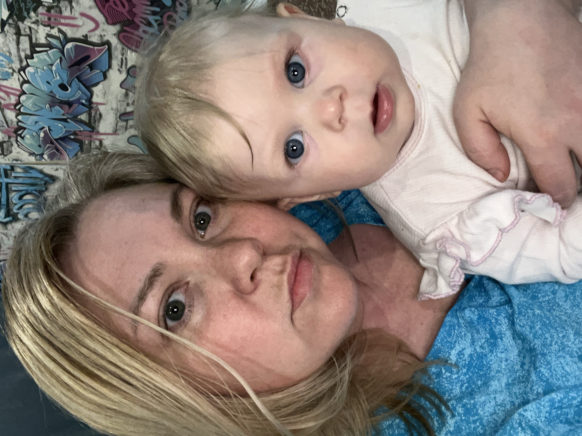 This image shows a selfie of a light-skinned woman with blonde hair and a baby with blue eyes. The woman has a serious expression and is lying down with the baby resting on her chest. The baby is wearing a light pink shirt, and the woman is wearing a blue shirt. There appears to be graffiti on the wall behind them. The location is unknown.