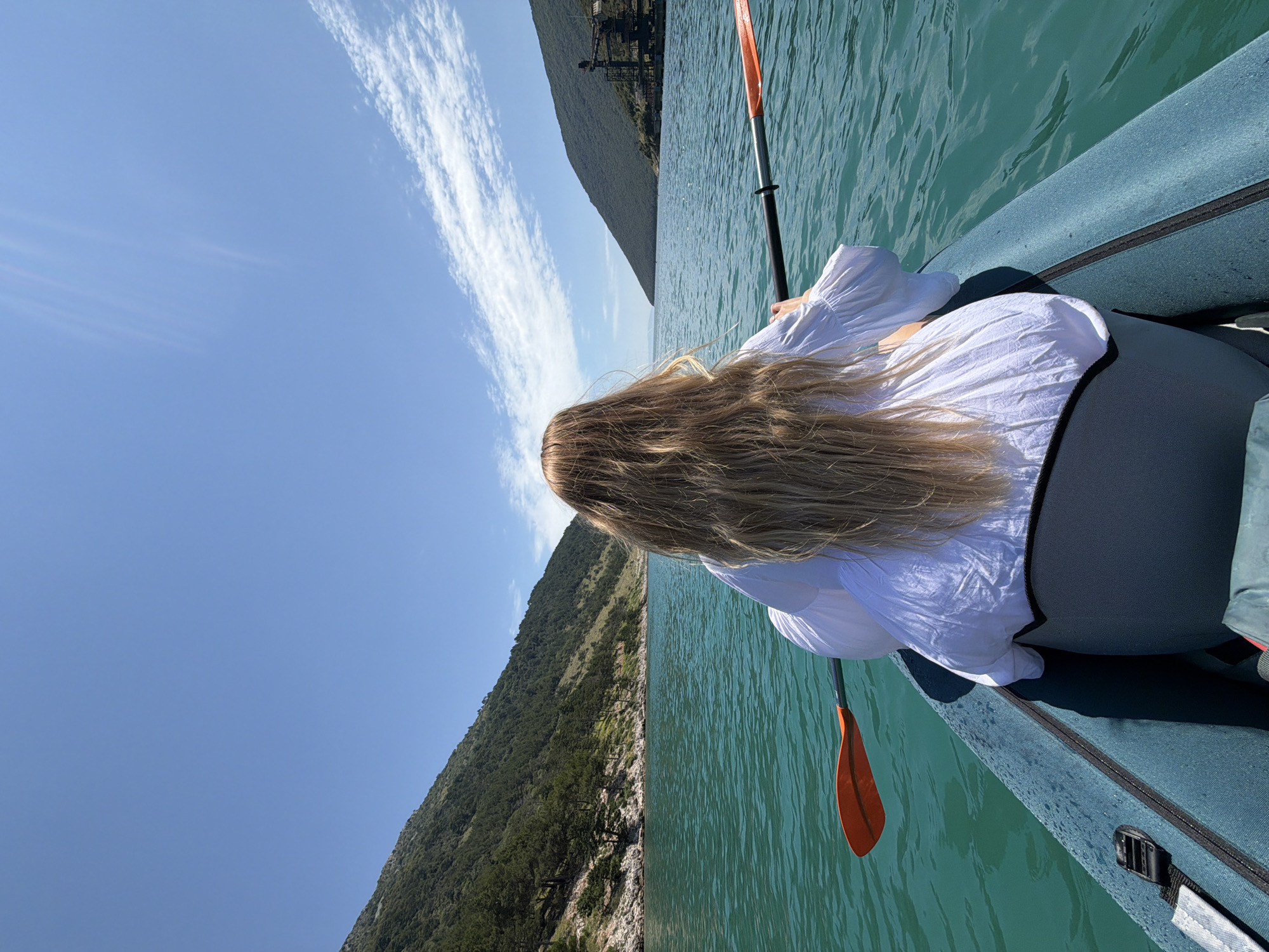 A first-person perspective shot shows a woman kayaking on a body of turquoise water. She has long, blonde hair and is wearing a white blouse. The woman is holding a black and orange paddle, with the orange end visible on the left side of the frame. The kayak is a dark blue inflatable boat. In the background, green, tree-covered mountains rise from the water on both sides, with a clear blue sky above, dotted with a few wispy white clouds. On the right side of the background, a distant industrial structure with cranes is visible near the shoreline.
