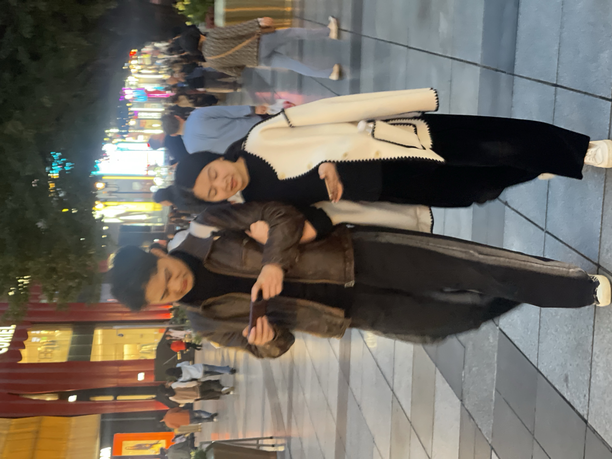 The image displays two East Asian individuals, a male and a female, walking arm-in-arm through a brightly illuminated urban pedestrian zone at night in Yuzhong District, China. The male, on the left, wears a dark turtleneck, a brown leather jacket, dark pants with a side stripe, and light sneakers. He holds a smartphone horizontally in both hands, actively viewing its screen. His right arm is linked with the female's left arm. The female, on the right, wears a black top, black wide-leg pants, and an open, cream-colored jacket with black trim. She holds a white cylindrical object in her right hand and wears light sneakers, looking slightly downward and to her right. The pavement consists of light and dark grey tiles arranged in a checkered pattern. In the background, numerous other individuals are visible, blurred due to movement. Tall commercial buildings line the street, featuring large illuminated windows and signs, including a partially visible "CURIEL" sign and distinctive red facade elements. The scene is well-lit by diverse artificial lights emanating from buildings and street infrastructure, casting yellow, blue, red, and pink hues. Dark tree foliage is discernible above the street level.