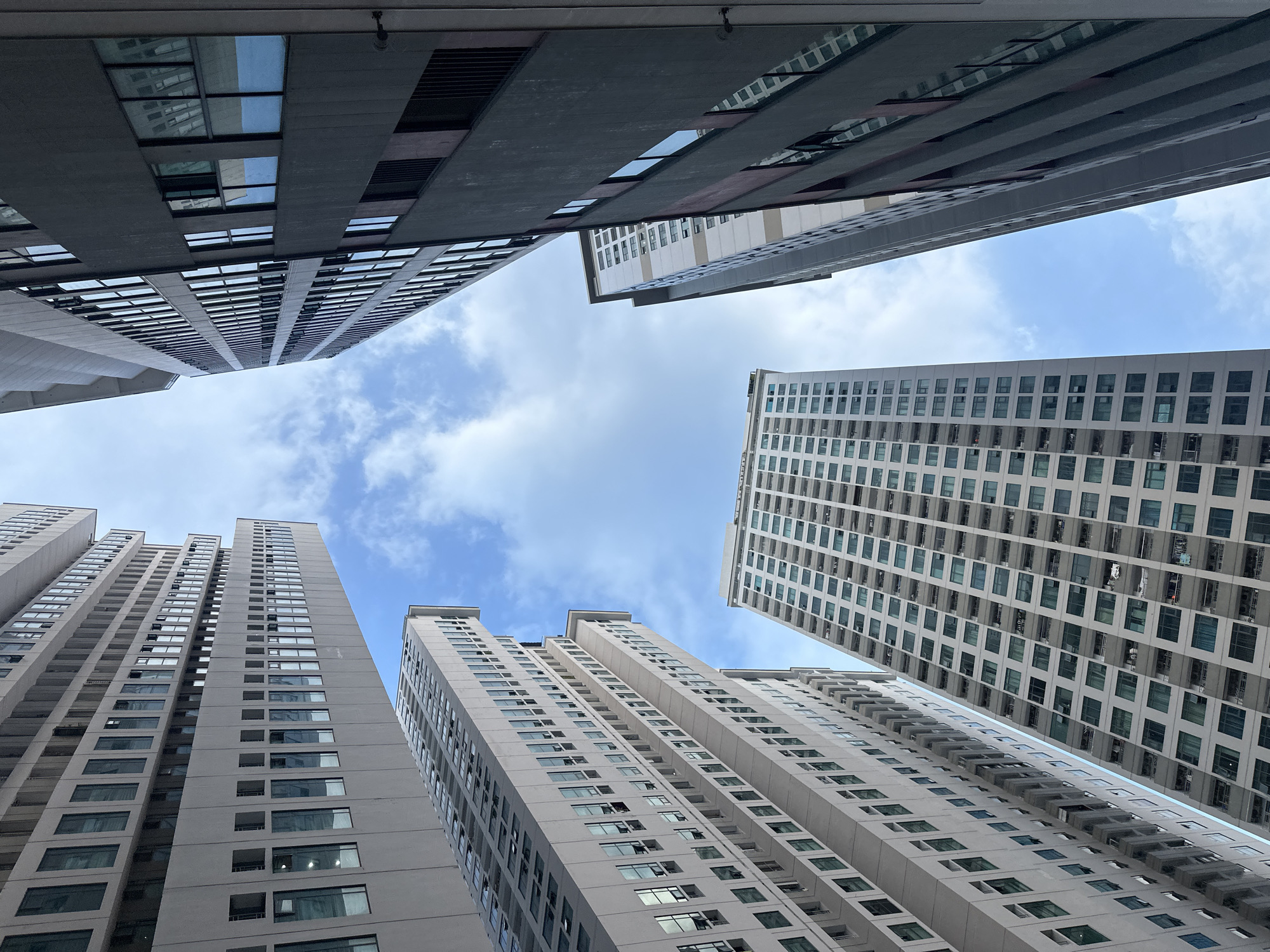 Looking up from the ground, the image shows several high-rise buildings in Bac Nha Trang Ward, Vietnam, angled against a blue sky with scattered white clouds. The buildings are predominantly gray and white, with numerous windows reflecting the sky. The perspective creates a sense of confinement, highlighting the density of the urban landscape. No people are visible in the frame.
