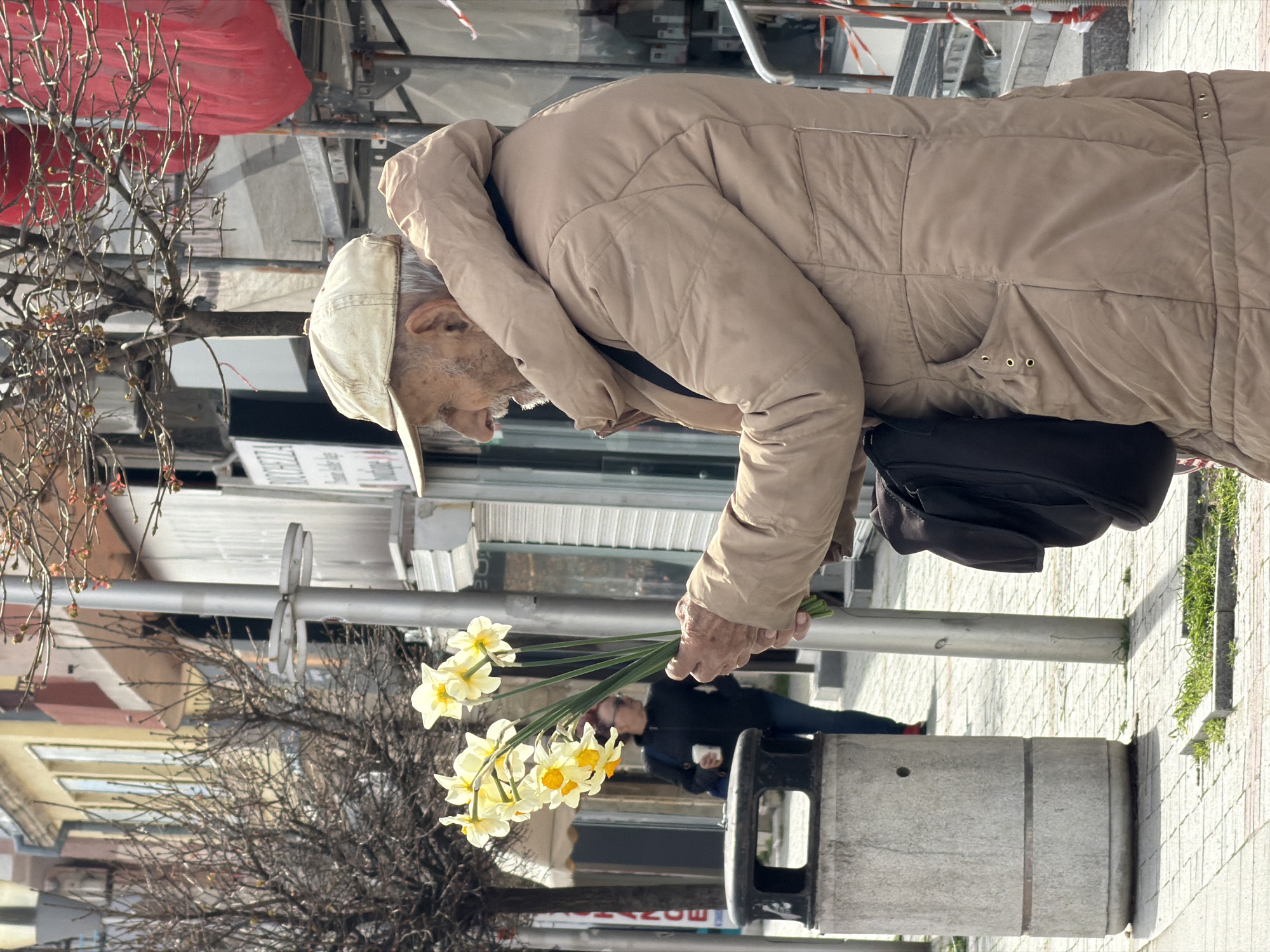 An elderly man, wearing a light-colored jacket and cap, holds a bouquet of daffodils in his right hand. A black shoulder bag hangs on his left side. He is standing on a street in Varna, Bulgaria. The street is paved with light-colored tiles. Behind him is a metal pole, a concrete trash receptacle and a person sitting on a bench. A bare tree is in the foreground. Several buildings are visible in the background.