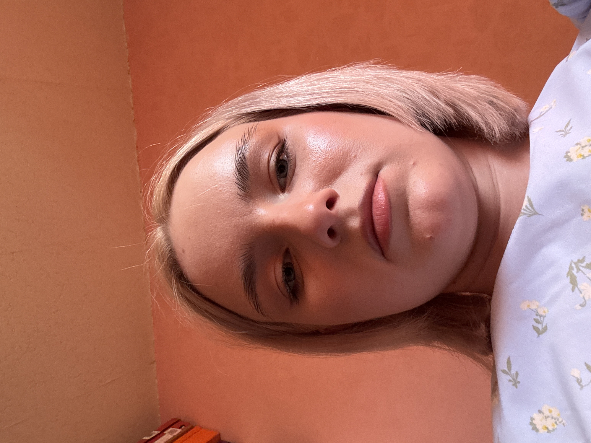 A close-up portrait captures a young woman, likely in her late teens or early twenties, positioned with her head tilted, looking directly towards the camera. Her fair skin appears dewy, highlighted by soft, warm lighting. She has blonde, bob-length hair and natural, well-defined eyebrows. Her lips are slightly parted, showing a natural tone or light lip tint. Her expression is neutral and calm.
She is wearing a light blue garment adorned with a delicate white and green floral pattern, which is visible on the right side of the frame. The background consists of two warm, muted orange/terracotta-colored walls meeting in a corner, suggesting an indoor setting. In the bottom-left corner, indistinct red and orange objects are partially visible. The activity is a posed portrait or selfie. No text is visible in the image. This scene takes place indoors in Saint Petersburg, Russia.