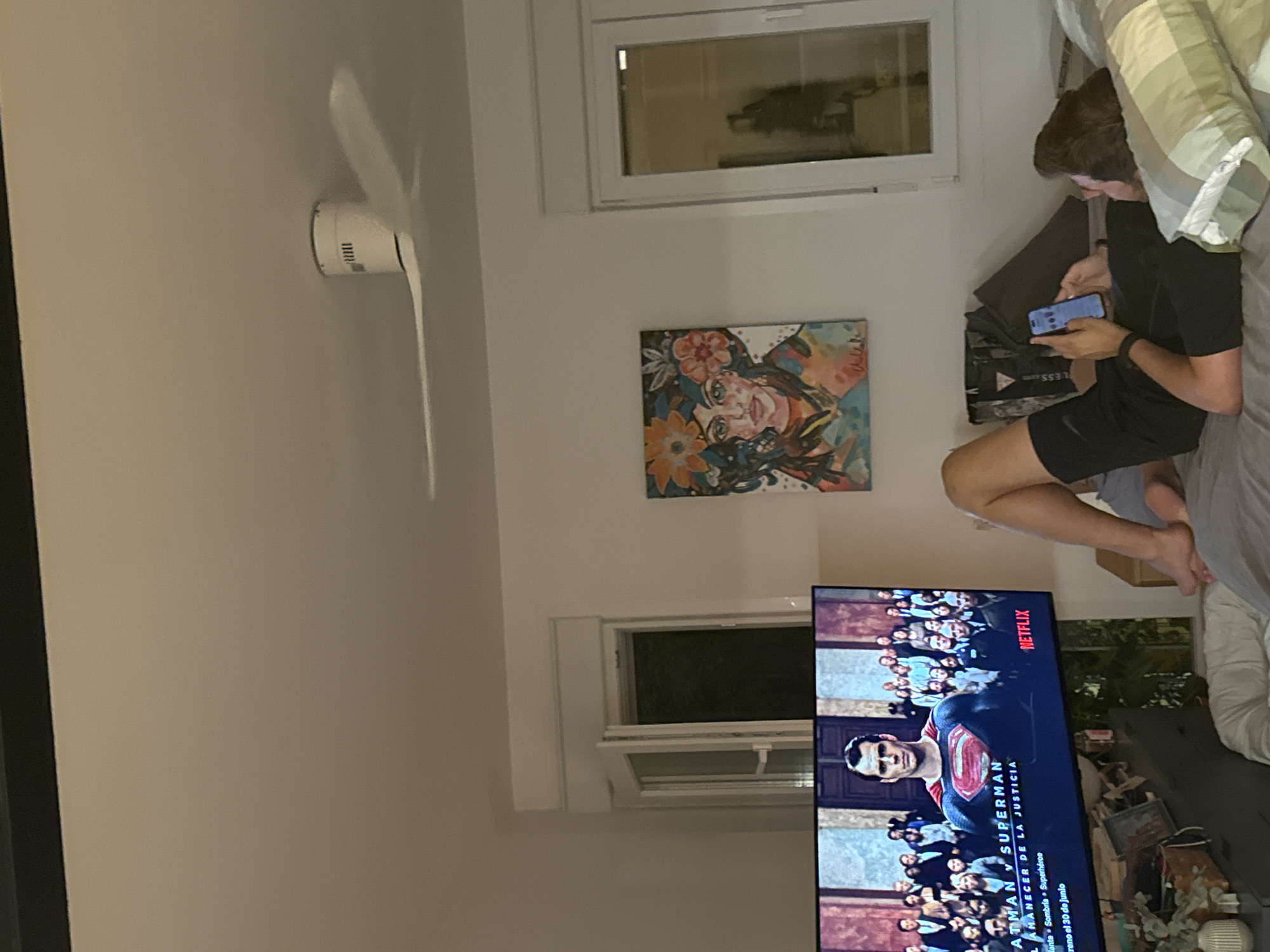 The image depicts an indoor scene, likely a bedroom, with a person lying on a bed looking at a smartphone. The room features a television displaying the movie "Batman v Superman: Dawn of Justice" with Spanish subtitles. A ceiling fan is visible above. Artwork of a woman with flowers in her hair hangs on the wall. There is a window with a view of what appears to be a balcony or exterior space. The overall impression is that of a casual, home entertainment setting. The image is not specific to Murcia, Spain; there are no identifiable landmarks or contextual clues to suggest this location.