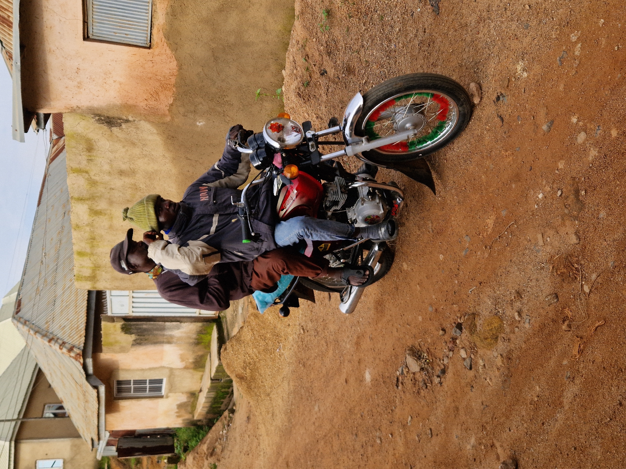 Two individuals are riding on a red motorcycle on a dirt road in the city of Jos, Nigeria. The person in front is driving the motorcycle and wearing a dark jacket with "NAIV" printed on the back, blue jeans, and black shoes. They have a green beanie on their head and appear to be holding onto the handlebars. The person behind the driver is wearing a dark long-sleeved shirt, brown pants, and black sandals. They are holding a light-colored bag or bundle in their arms and have a dark baseball cap on.
The environment consists of rustic, earthen-colored buildings with corrugated metal roofs, suggesting a residential area. Piles of sand or soil are visible on the side of the road, along with some sparse vegetation. The ground is uneven and covered with reddish-brown dirt, characteristic of many roads in Nigeria. The sky is overcast, indicating a cloudy day. The image captures a candid moment of transportation and daily life in Jos.