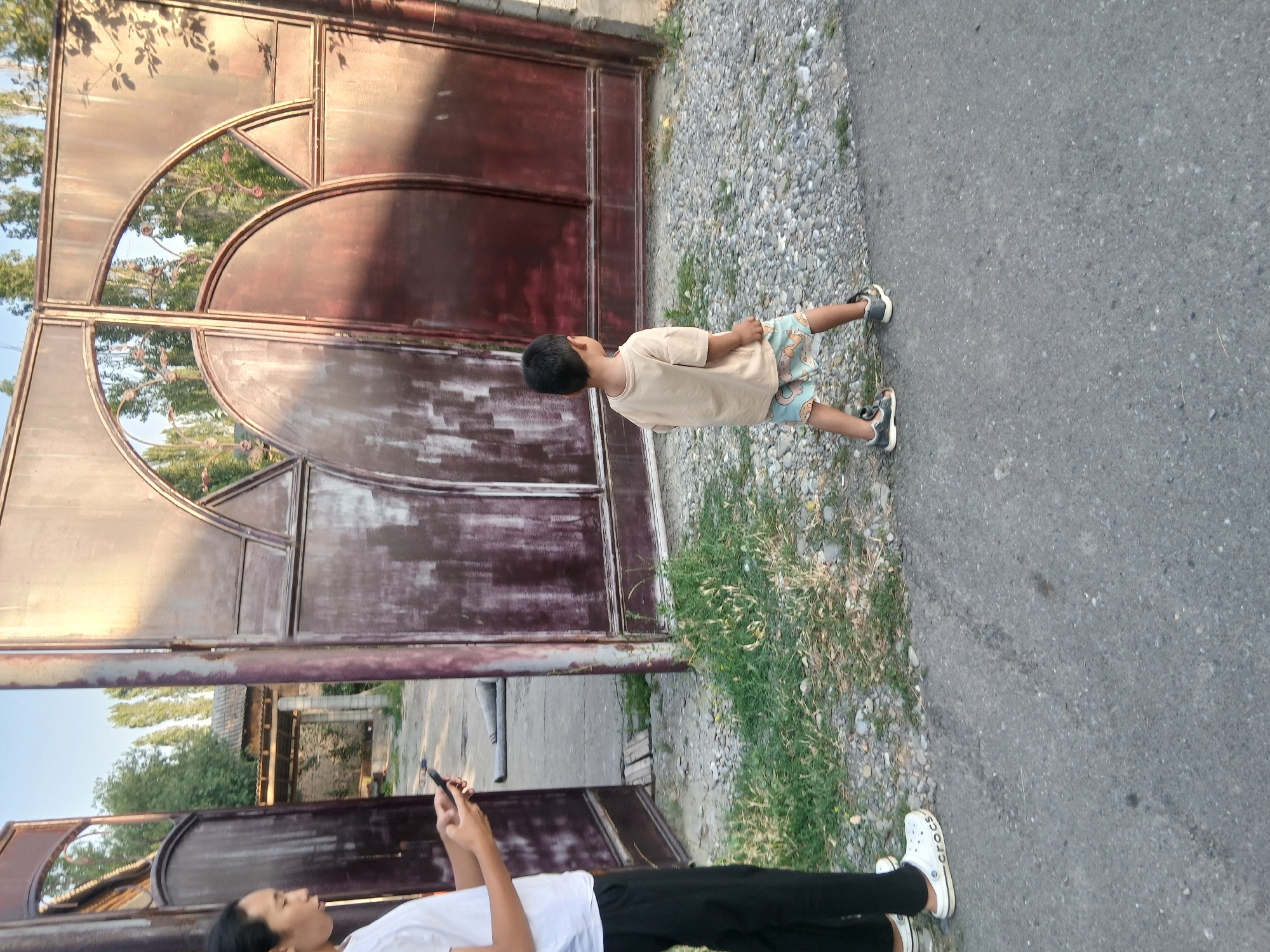 The image shows two people standing near a large, rustic metal gate in Тайгараев, Kyrgyzstan. On the left, a person is standing with their back to the camera, wearing a white t-shirt and black pants, and holding a black smartphone. White Crocs are visible on their feet. To the right, a young child, seen from behind, is walking away from the camera towards the gate. The child is wearing a light tan t-shirt and patterned shorts, with dark sneakers on their feet. The gate is made of dark metal with ornate circular and triangular patterns. Behind the gate, trees and buildings are visible. The ground in front of the gate is a mix of gravel and dry grass. The scene appears to be outdoors, possibly in a residential area, with a paved surface in the foreground.