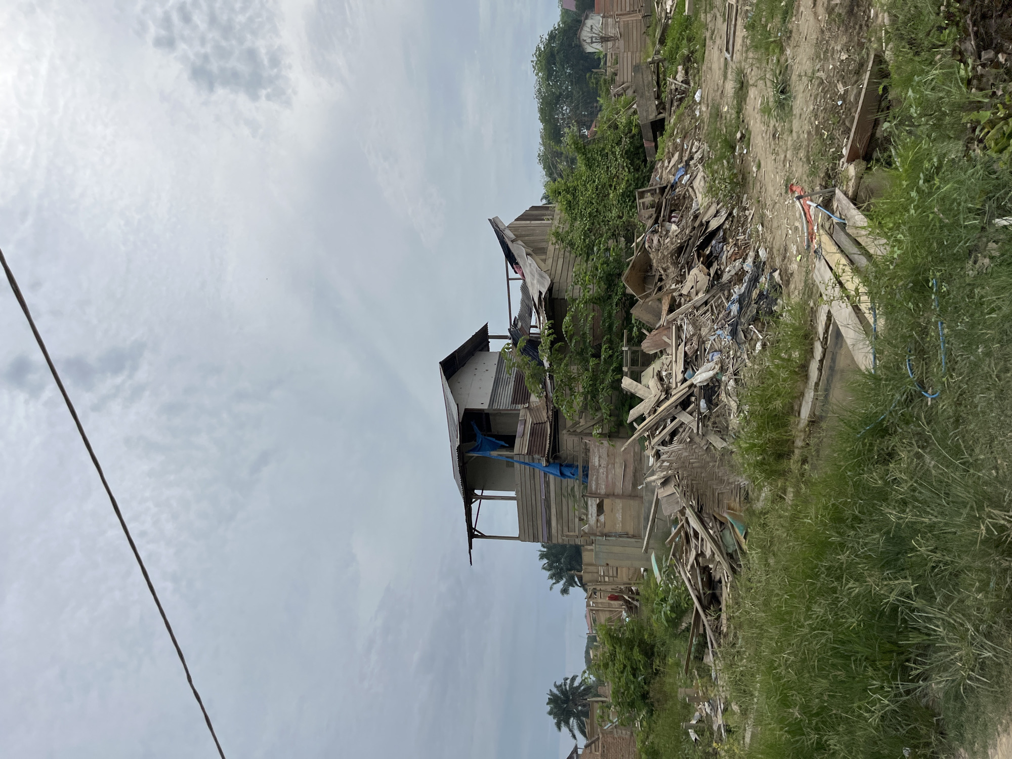 In Bundar, Indonesia, an overcast sky looms over a scene of destruction and disarray. The remains of a structure, likely a house, are partially visible amidst a landscape littered with debris. Piles of wood, corrugated metal sheets, plastic sheeting, and other construction materials are scattered haphazardly across the ground. Overgrown grass and vegetation are encroaching on the scene, suggesting a period of neglect or abandonment. The skeletal remains of the building, with its exposed wooden framework and partially intact roof, speak of a recent demolition or severe damage. The overall impression is one of a community in a state of rebuilding or recovery, with the remnants of past structures serving as stark reminders of past events. The presence of palm trees in the background hints at a tropical or subtropical environment.