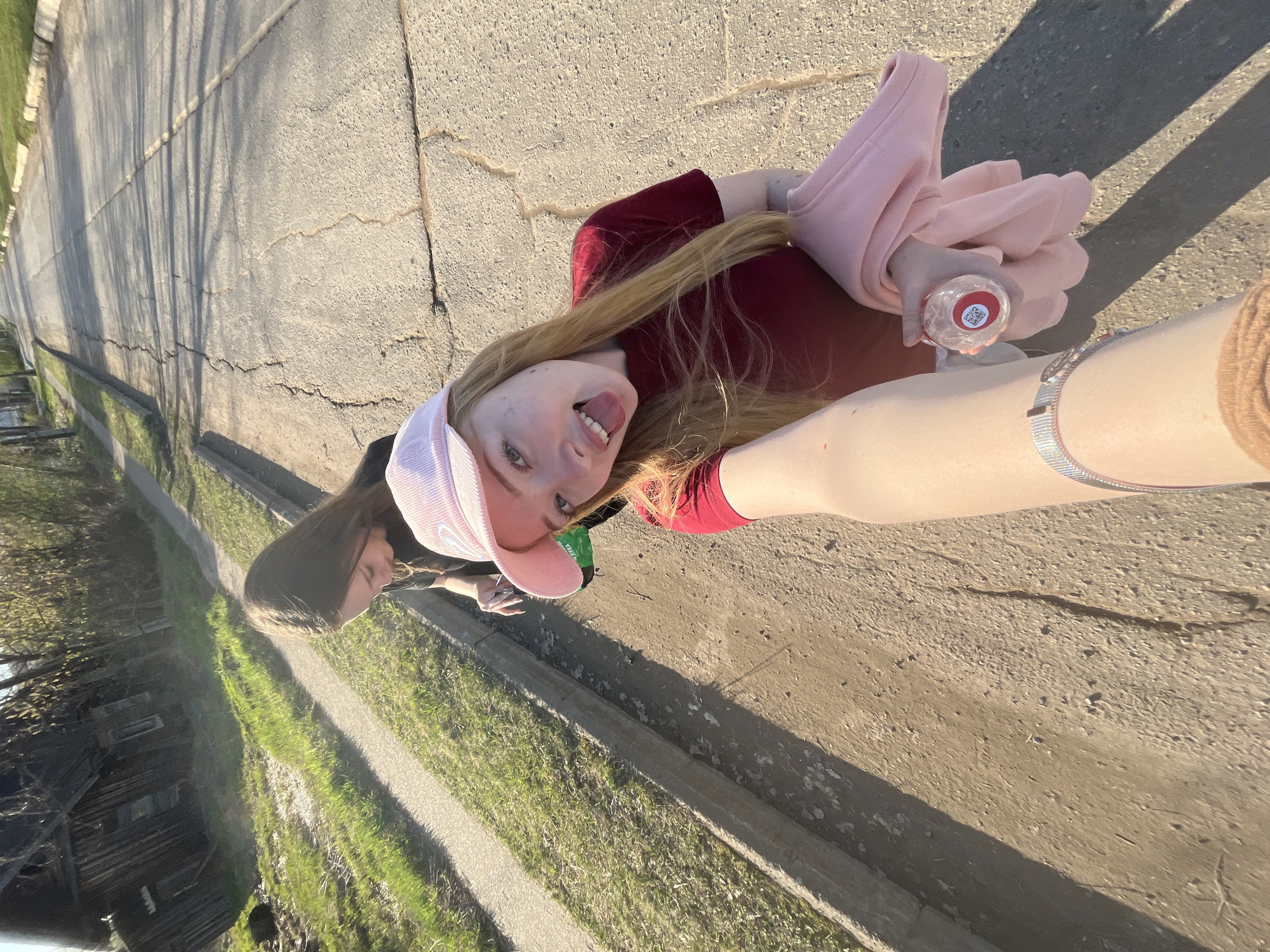 Two people are standing on a cracked concrete road in Сосново, Russia. One person is holding the camera, wearing a pink baseball cap, a maroon shirt, and a silver watch on their left wrist. They are holding a clear water bottle and a pink sweatshirt. The other person is standing behind them. The road is bordered by a grassy area with a concrete curb. Trees and a small wooden structure are visible in the background.