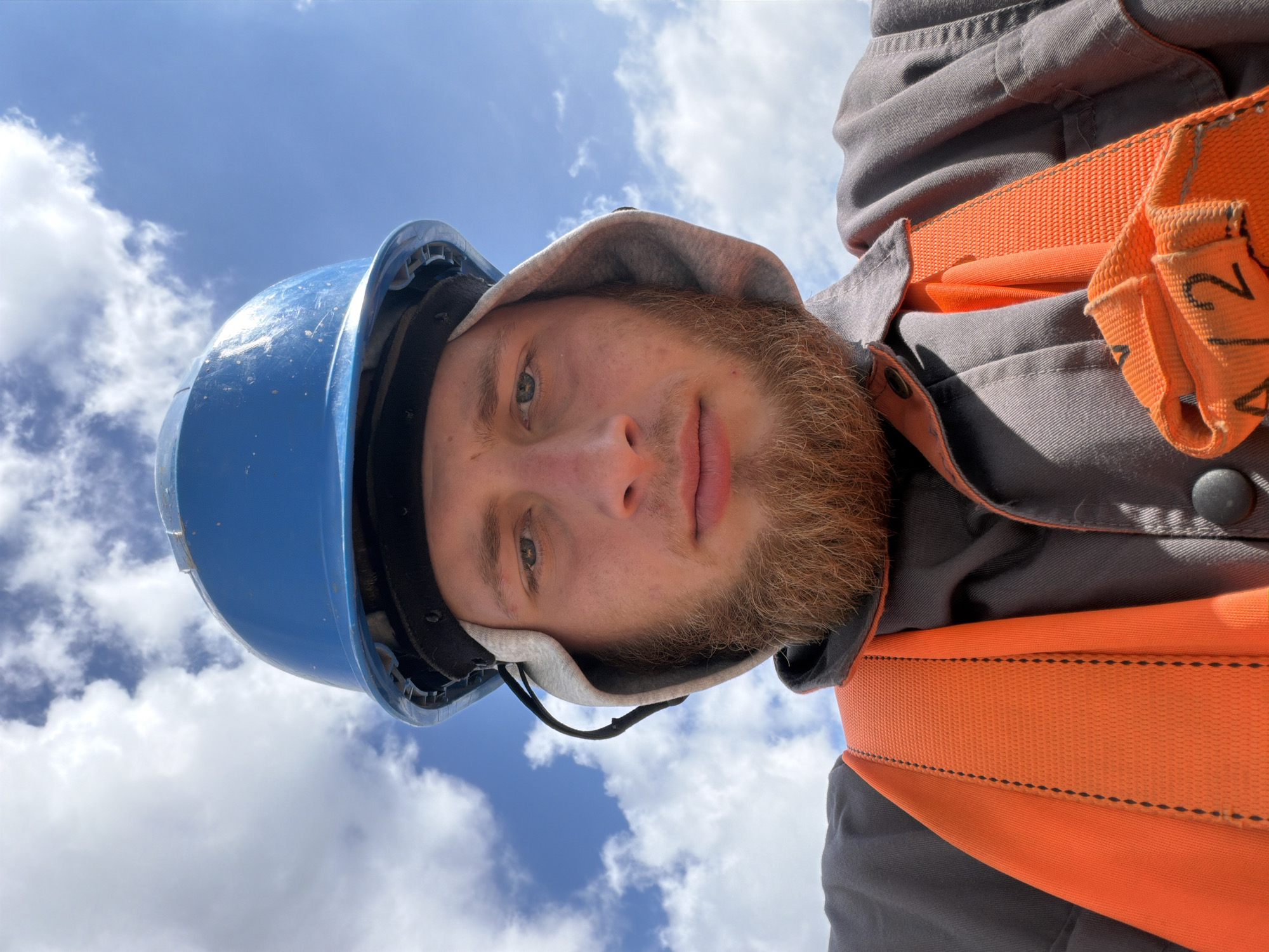 A close-up, low-angle, selfie-style photograph depicts a young Caucasian male with light skin and a full reddish-brown beard. He is wearing a blue hard hat, a grey work jacket, and an orange safety harness over the jacket. The man is looking directly at the camera with a neutral expression. The background consists of a bright blue sky with scattered white clouds. The setting appears to be outdoors, possibly on a construction site or elevated platform, given the safety gear. There is no specific indication of the location being in the city of Stróża, Poland, within the image itself. The lighting suggests it is daytime.