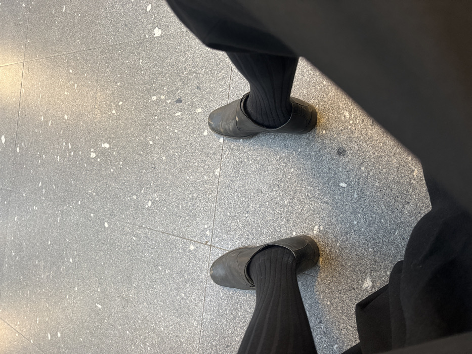 A low-angle, downward-looking image captures the lower extremities of one individual on a tiled floor. The person is wearing black trousers, black ribbed socks, and a pair of worn, dark-colored, slip-on shoes with visible scuff marks. The left foot is positioned slightly forward and to the left of the frame's center, while the right foot is to the right. The floor consists of light gray rectangular tiles with irregular white speckles, indicative of terrazzo or a similar composite material. Grout lines delineate the tile pattern. Reflections of overhead light sources are visible on the polished tile surface, particularly around the shoes. The individual appears to be seated or standing stationary. No specific identifying markers for a precise location within Guimarães, Portugal, are discernible from the visual information provided.