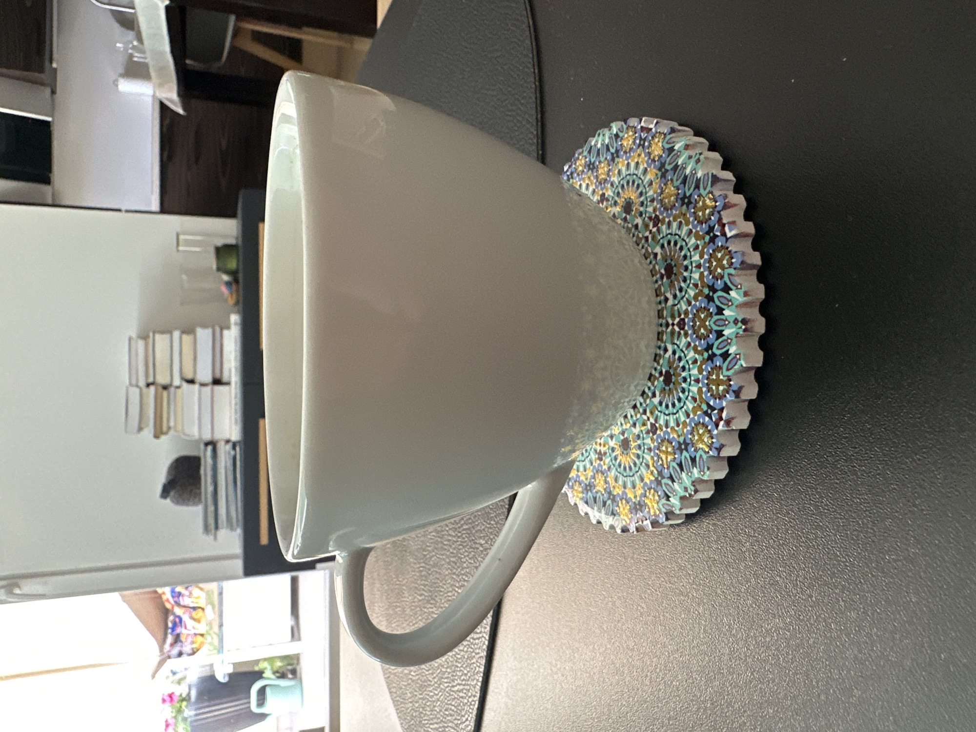 This media file depicts a close-up, rotated view of a white ceramic mug resting on a decorative coaster. The mug is large and appears to be a coffee or tea cup, with a curved handle visible. The coaster is round with a serrated edge and features an intricate, colorful mosaic pattern in shades of blue, yellow, green, and brown, reminiscent of Moroccan tiles. The mug and coaster are placed on a dark, textured surface, possibly a table.
In the background, out of focus, are hints of an indoor setting. To the left, a stack of books or similar items is visible, with a small, dark object that might be a figurine or a small electronic device. Further left and out of focus, there are glimpses of furniture and what appears to be a window with reflections of plants and possibly colorful objects, suggesting a domestic environment. The lighting indicates it is daytime, likely indoors, with natural light entering from a window.
There is no visible text in the image. The overall impression is a still life composition highlighting the textures and patterns of the mug and coaster in a domestic setting. The location context of Warsaw, Poland, does not offer specific visual cues within this particular crop.