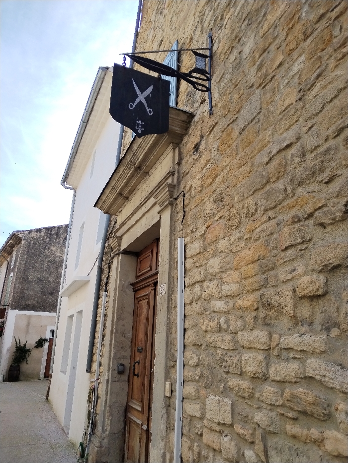 A stone building features a dark flag with white scissors hanging above an ornate doorway. The door is wooden with an address number visible. A metal sign with scissors extends from the building, suggesting a tailor or barber shop. A narrow street runs alongside the building, revealing a glimpse of another structure further down the street. The sky is partly cloudy.