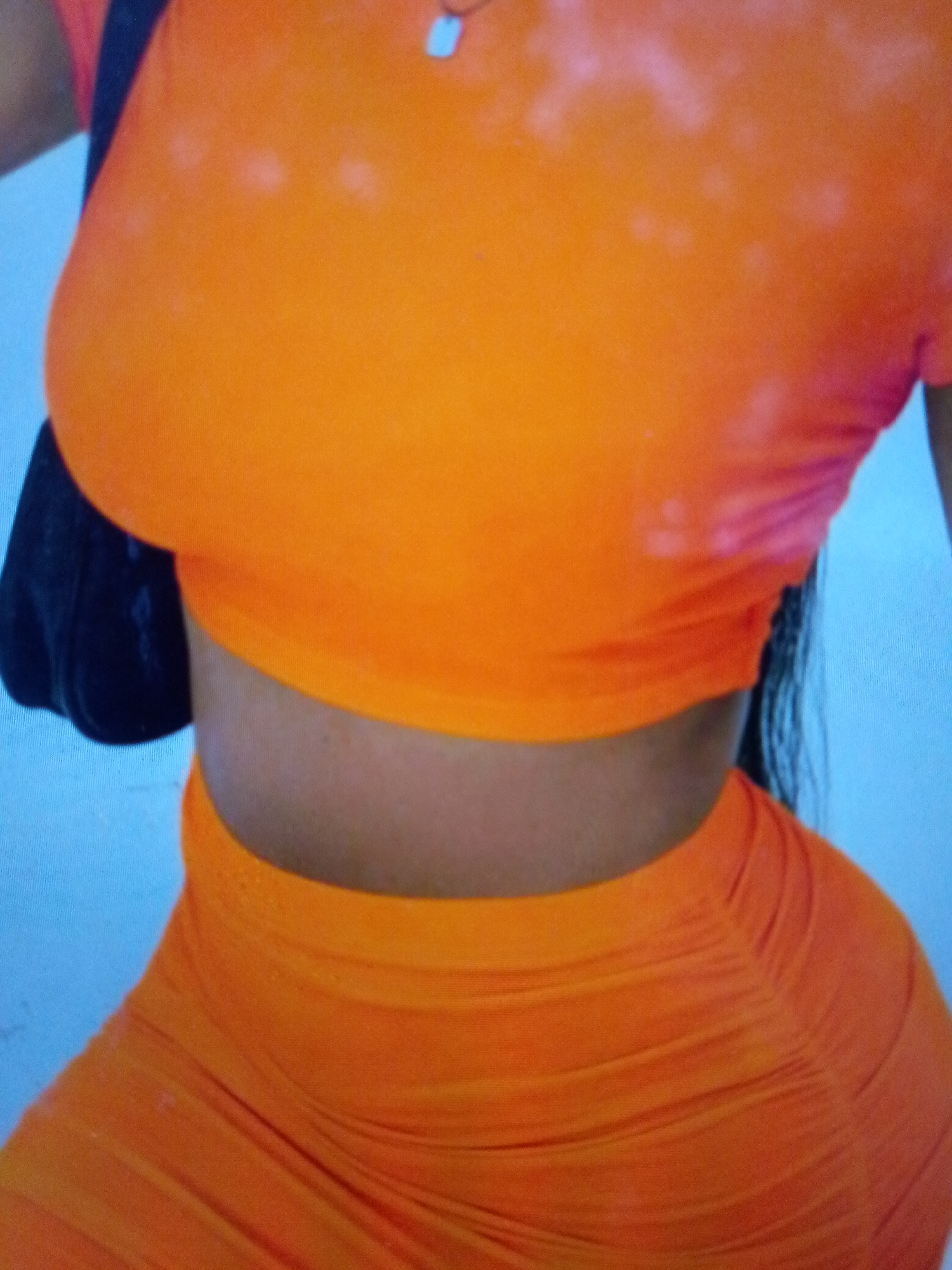 A torso-level view of a person wearing a matching orange cropped top and skirt. The top has bleached patches, and a black bag strap is visible on the left shoulder. The skirt is ruched. The background is light blue.