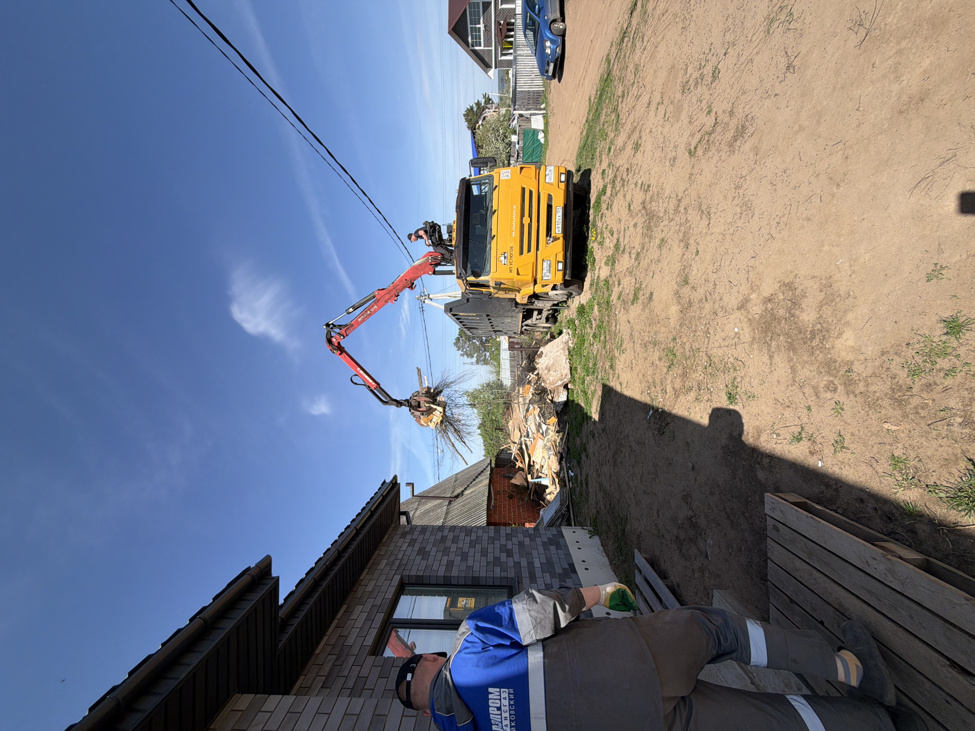 The scene depicts a construction or demolition site in Chaikovsky, Russia, evident from the Cyrillic text on the worker's uniform ("ГАЗПРОМ ТРАНСГАЗ ЧАЙКОВСКИЙ"). A yellow KAMAZ truck with a red crane arm is positioned near a pile of debris, apparently remnants of a demolished structure. The crane arm is actively lifting and possibly moving the debris.
A building constructed of gray brick is in the foreground. A man wearing gray work overalls with blue accents, along with reflective stripes and a baseball cap, stands in the lower part of the frame, his body partially obscured. The background includes a blue sky with power lines stretching across it, along with other buildings and vegetation in the distance. The ground consists of dirt or gravel. A blue car is in the background.