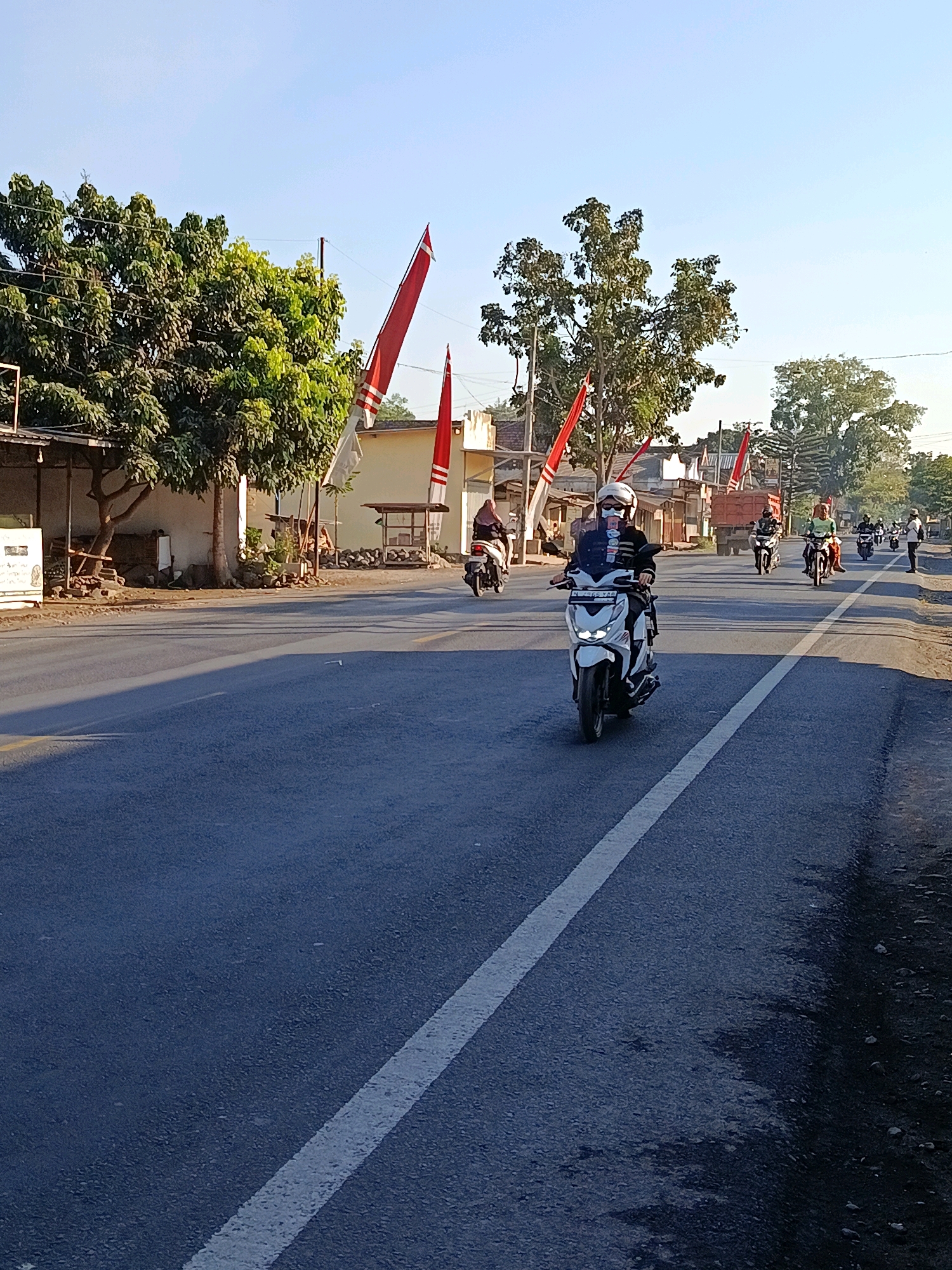 The image depicts a street scene in Tempeh Lor, Indonesia. Several people are riding motorcycles on a paved road with a white line marking the lane. In the foreground, a person on a white scooter is wearing a helmet and a face mask. To the left of the road, there are buildings and trees, with red and white flags displayed. In the background, more motorcycles and a truck are visible. The sky is clear and blue, indicating daylight.