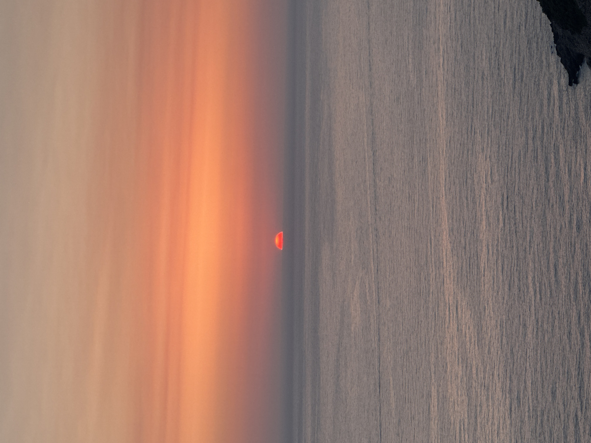 A sunset is depicted over the Adriatic Sea, with the sun appearing as a small, reddish-orange semi-circle on the horizon. The sky above is a gradient of soft oranges and pinks, transitioning to a muted grey towards the top. The sea below is a calm, greyish-blue, with subtle ripples reflecting the sky's colors. In the lower right corner, a dark, rocky outcrop juts into the water. The scene captures the serene beauty of twilight.