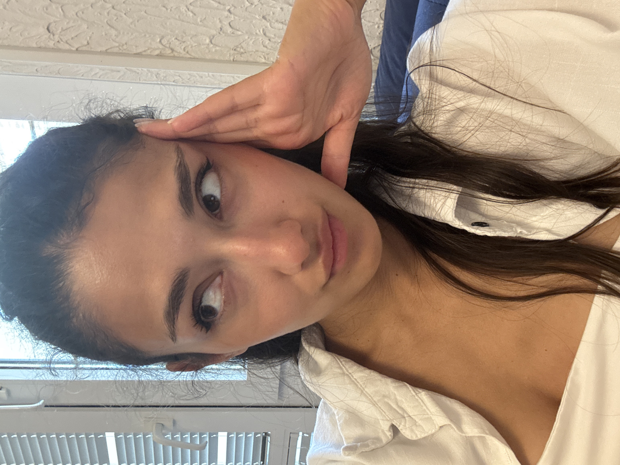 A young woman with long, dark hair pulled back partially, gazes to her left, her left hand resting on her temple. She is wearing a white shirt, open at the collar, revealing her neck and upper chest. The background includes a window with blinds on the left and a textured wall on the right. A dark blue surface, possibly a couch, is visible in the lower right corner. The scene appears to be indoors, likely in an apartment or hotel room. This image was taken in Sutomore, Montenegro.