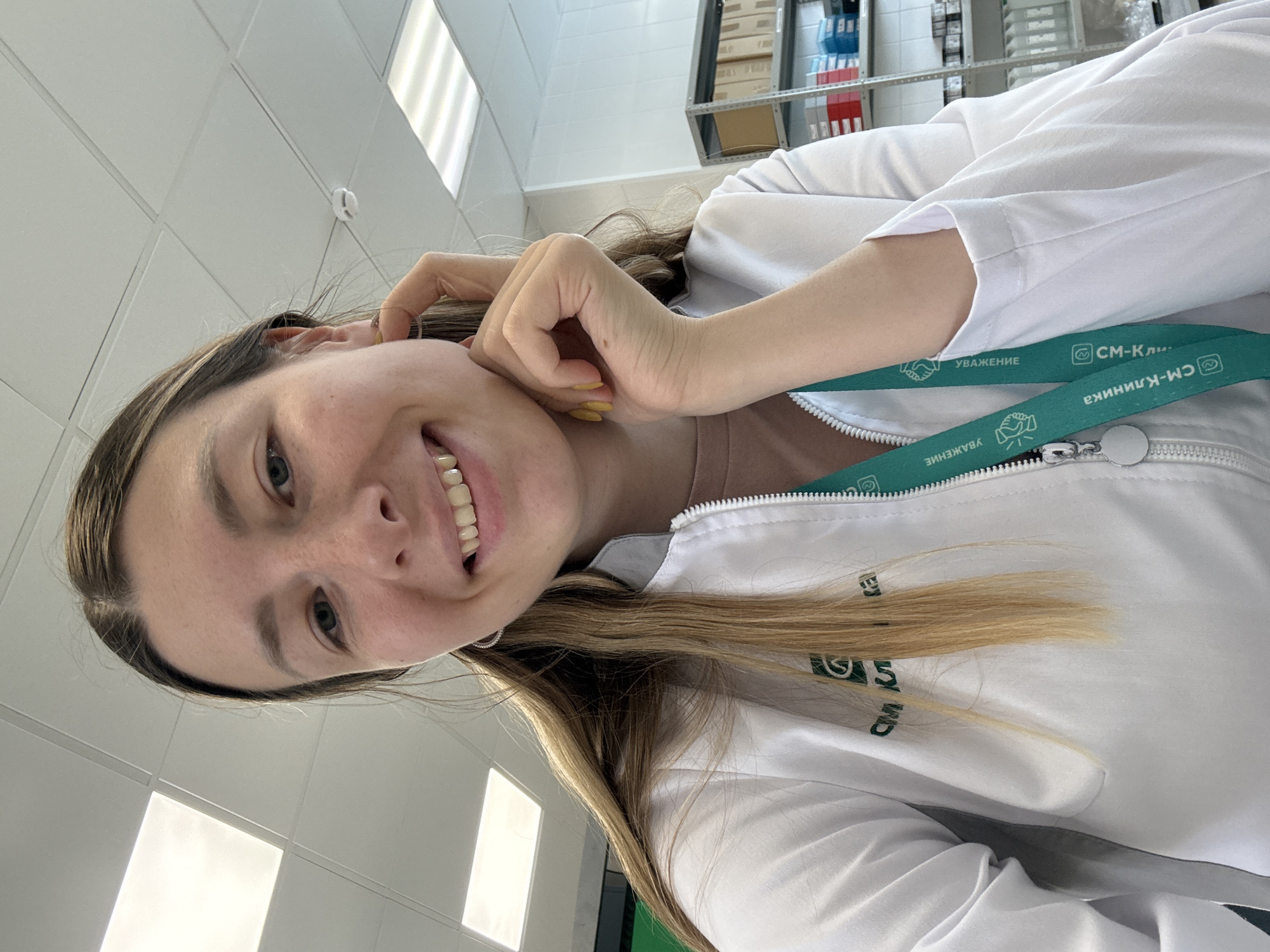 A young woman with long, blonde hair and blue eyes smiles as she looks at the camera. She is wearing a white lab coat with a green lanyard. Her right hand is resting on her chin. The background appears to be an indoor setting with fluorescent lighting and shelves stocked with medical supplies. The environment suggests a clinic or laboratory. There is no visual information that places the scene in Moscow, Russia.