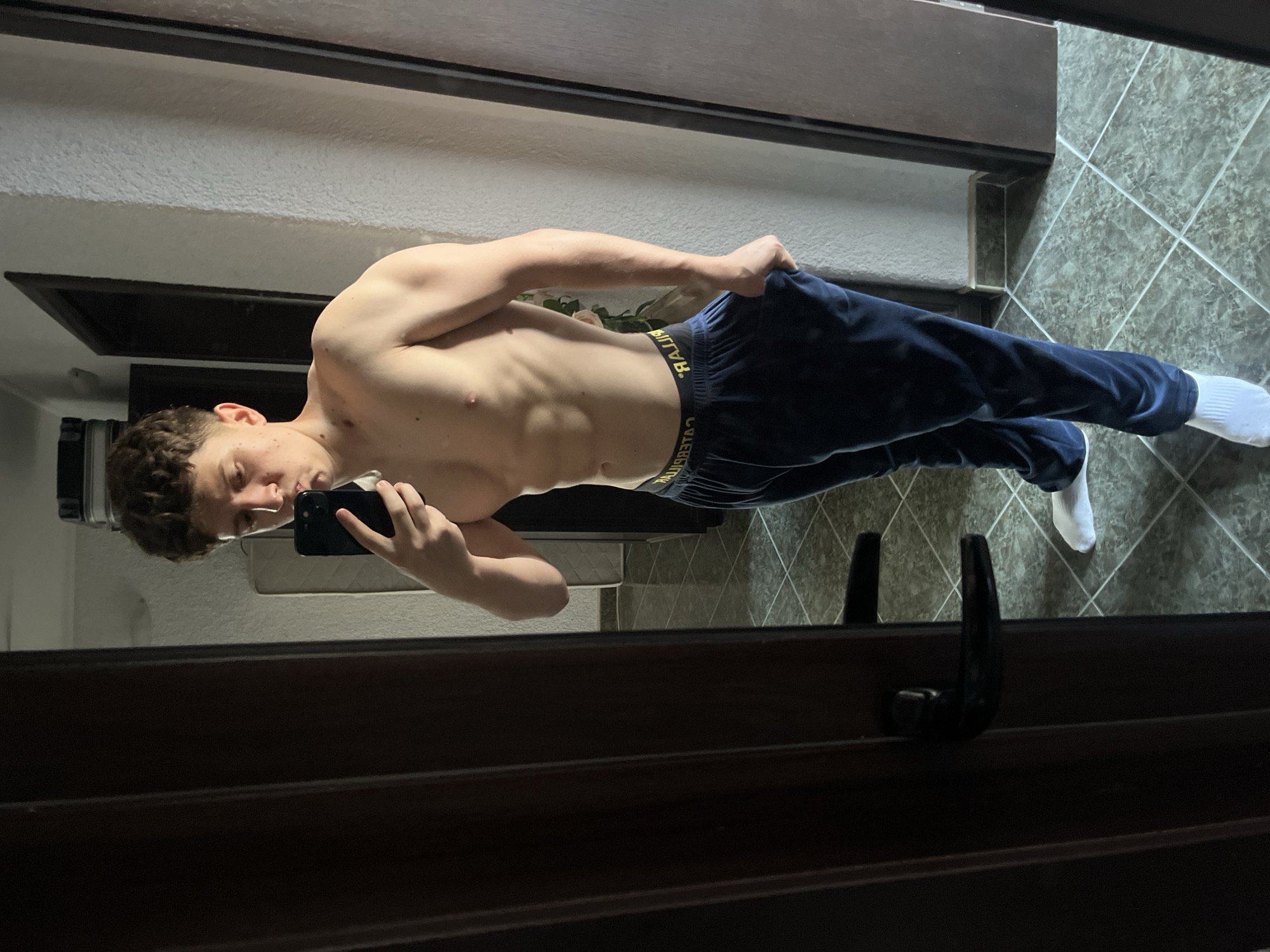 A young male individual is captured in a full-length mirror selfie, presumably located in Zlatograd, Bulgaria. The subject is shirtless, displaying a lean and muscular physique with defined abdominal muscles and developed upper body musculature. He has short, curly brown hair and appears to be looking down at a black smartphone held in his left hand, which is positioned for taking the photograph. His right hand is partially inserted into the pocket of his dark blue athletic pants, which appear to be made of a soft material like velvet or fleece. The yellow and black waistband of "CATERPILLAR" branded underwear is visible above the pants. He is wearing white ankle socks. The reflection reveals an indoor setting with a dark wooden door featuring a black handle on the left side of the frame, and a light-colored, textured wall on the right. The floor is covered with grey-green tiled squares arranged in a diagonal pattern. Faintly visible in the distant reflection are what appears to be a mattress or furniture item and another dark door frame.