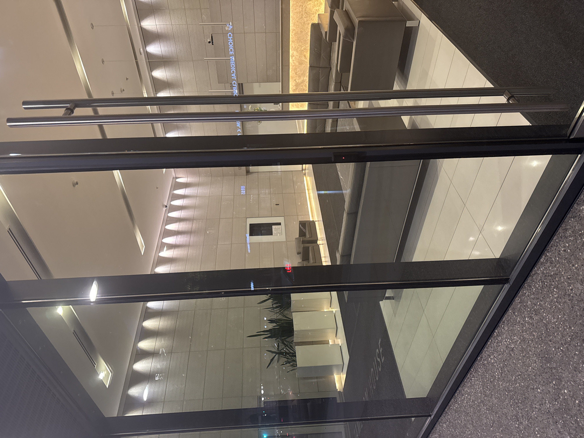 This image depicts the entrance to a modern building lobby. The view is through a glass facade with large, silver double doors in the foreground. The lobby itself is well-lit with contemporary architectural lighting.
Inside, a reception area is visible with a sleek, illuminated counter. Behind the counter, a sign reads "CHOICE MEDICAL CLINIC". Soft seating, including a grey leather sectional sofa, is arranged in a waiting area. The walls are finished with large, light-colored stone tiles, and the floor is a combination of white tiled areas and dark carpeting. Three tall white planters with green foliage are positioned to the left of the entrance, and a mat on the floor inside the entrance has the word "HOUSE" visible in white lettering. The scene suggests a professional and clean medical office environment. The presence of "CHOICE MEDICAL CLINIC" strongly suggests this location is in Vancouver, Canada, as this is a known clinic in that city.