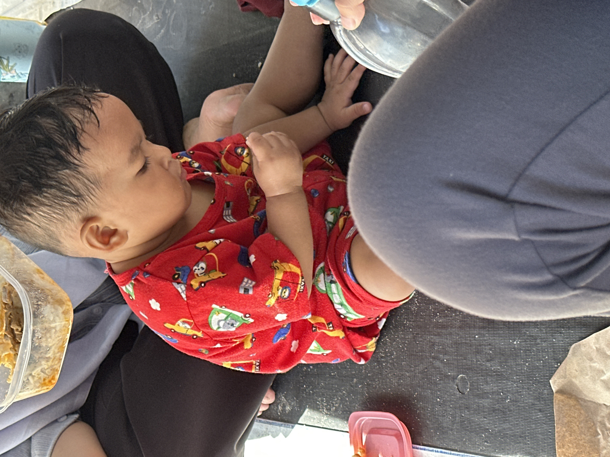 A baby in a red shirt with vehicle designs is lying down. The baby is being fed from a metal bowl by a person wearing grey clothing. A clear plastic container of food and a brown paper bag are on the surface. The baby is lying on a black surface. The location is Lhokseumawe, Indonesia.