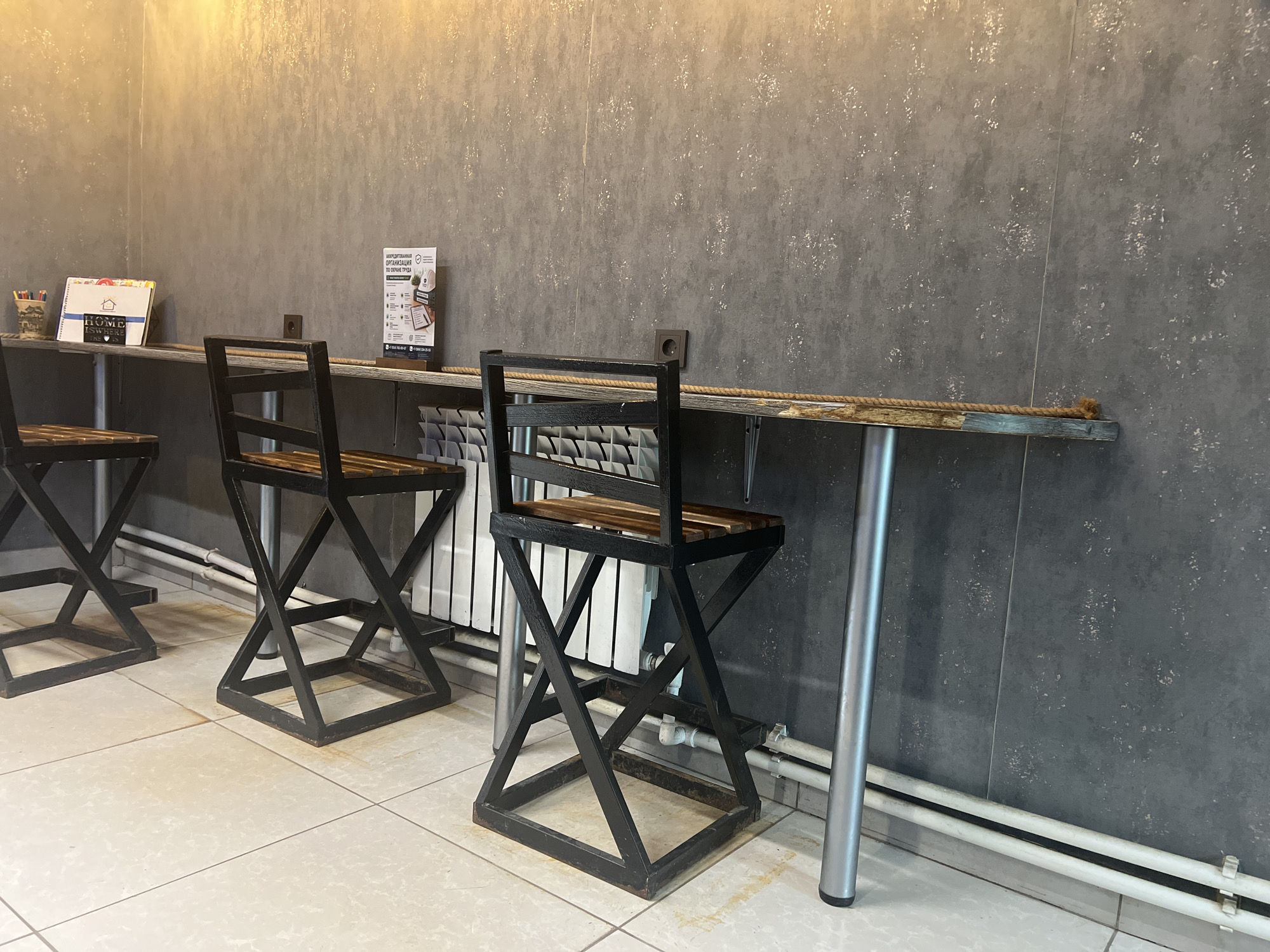 The image depicts a row of three black metal bar stools with wooden seats, positioned against a gray textured wall. The stools are arranged in front of a narrow, dark wooden counter, which is supported by metal legs and a bracket. The counter is positioned at a high level, suitable for bar seating. On the left side of the counter, a metal cup holds pens and pencils, and a small brochure or book with the word "HOME" visible is also present. To the right of these items, a white and red informational stand is displayed on the counter. Behind the stools, a white radiator or heating system is visible against the wall. The floor is tiled with light-colored square tiles. A thick rope is draped across the counter. The overall scene suggests an indoor seating area, possibly in a cafe, restaurant, or public space. The location is identified as Yuzhno-Sakhalinsk, Russia, based on the provided context. There are no people visible in the image, and no actions or interactions are taking place. The environment appears to be clean and orderly.