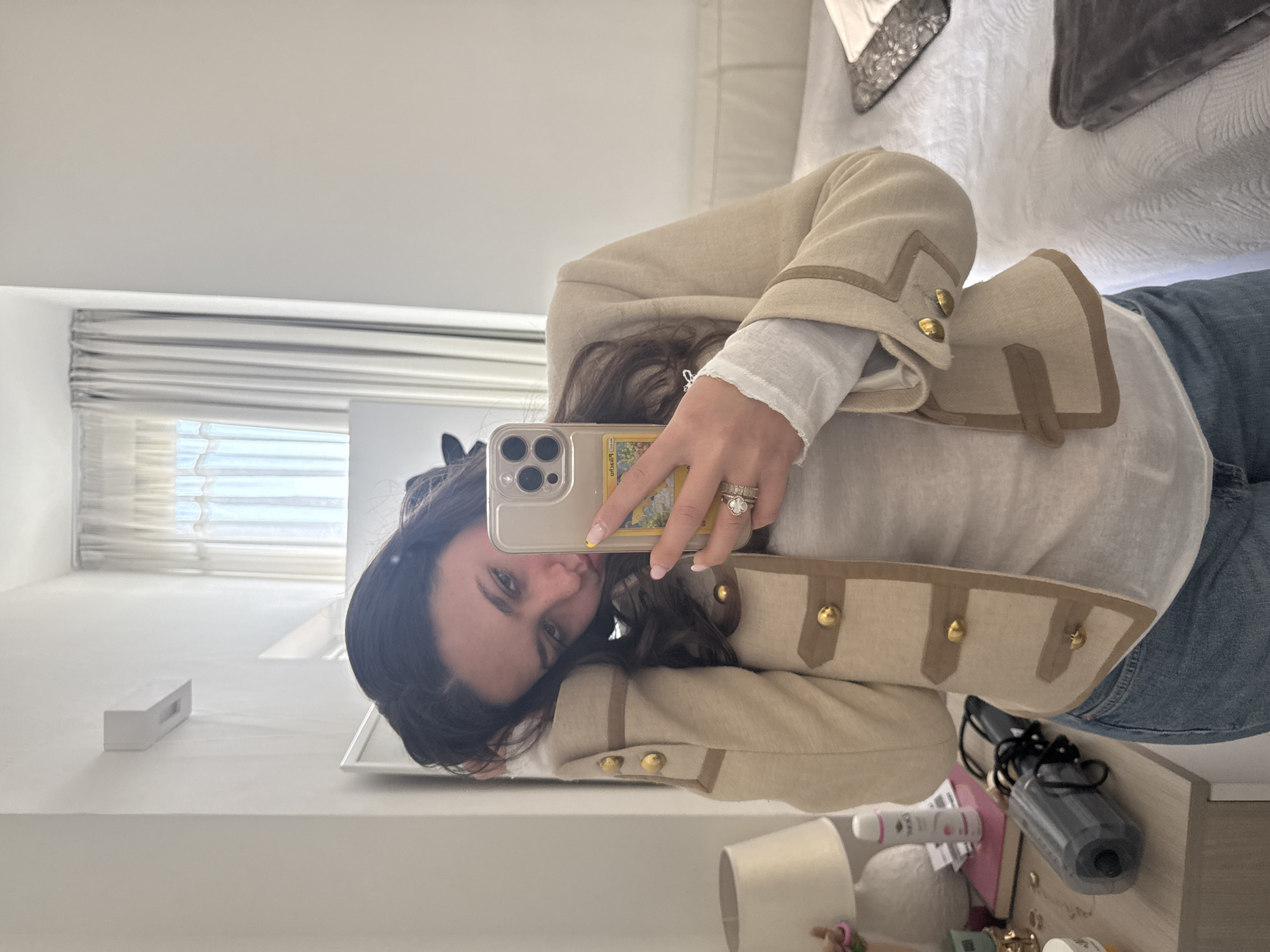A young woman is captured in a mirror selfie, with the image rotated 90 degrees counter-clockwise. She has long, dark wavy hair and looks towards the camera with a neutral expression. She wears a cream or beige military-style jacket featuring brown trim and gold buttons, layered over a white long-sleeved top, and blue jeans. Her left hand, holding a light-colored iPhone with a clear case, displays multiple rings on her ring finger, including a prominent cluster ring. A Pokémon card, likely depicting Pikachu, is visibly tucked into the phone case, and her fingernails are painted with light pink and a touch of yellow polish.
The setting is an indoor room, likely a bedroom or dressing area, in Milan, Italy. Natural daylight streams from a window with white horizontal blinds in the background. The room features light-colored walls. In the foreground, a white dresser or table is cluttered with various personal items, including a white rectangular speaker, a black hair dryer, a white cylindrical container, a bottle with "Dove" text, and a small white lamp with a beige shade. A portion of a bed with a white textured comforter and a grey throw blanket is visible in the upper part of the frame. The scene conveys a casual, everyday moment.