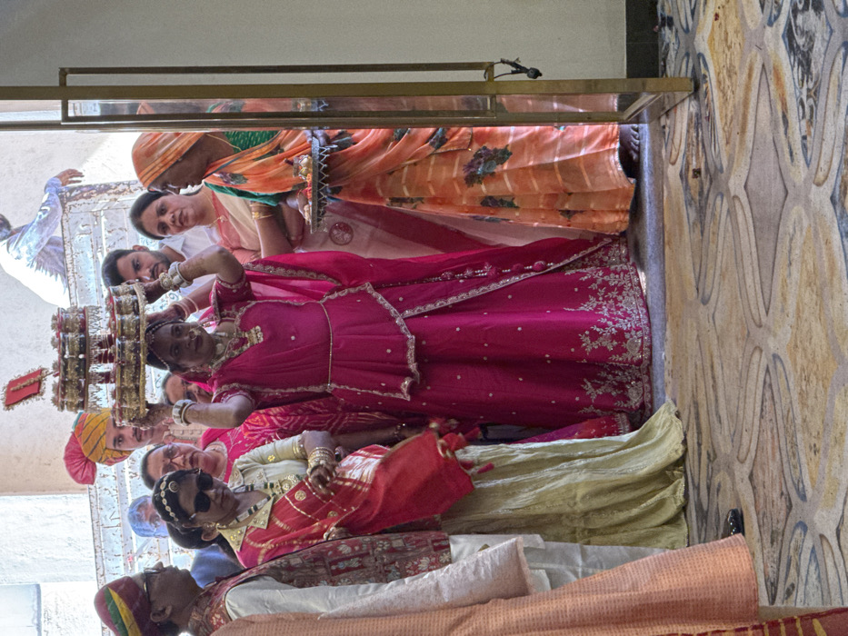 A group of people stand inside a building. Many are in bright, traditional Indian clothing. One woman wears a vibrant pink outfit with silver embroidery and holds a tiered, decorative structure above her head. Another individual sports sunglasses and a gold garment. Others wear colorful turbans and sarees. The backdrop includes an ornate wall and architectural details, suggesting a festive or ceremonial setting within a historic building.
