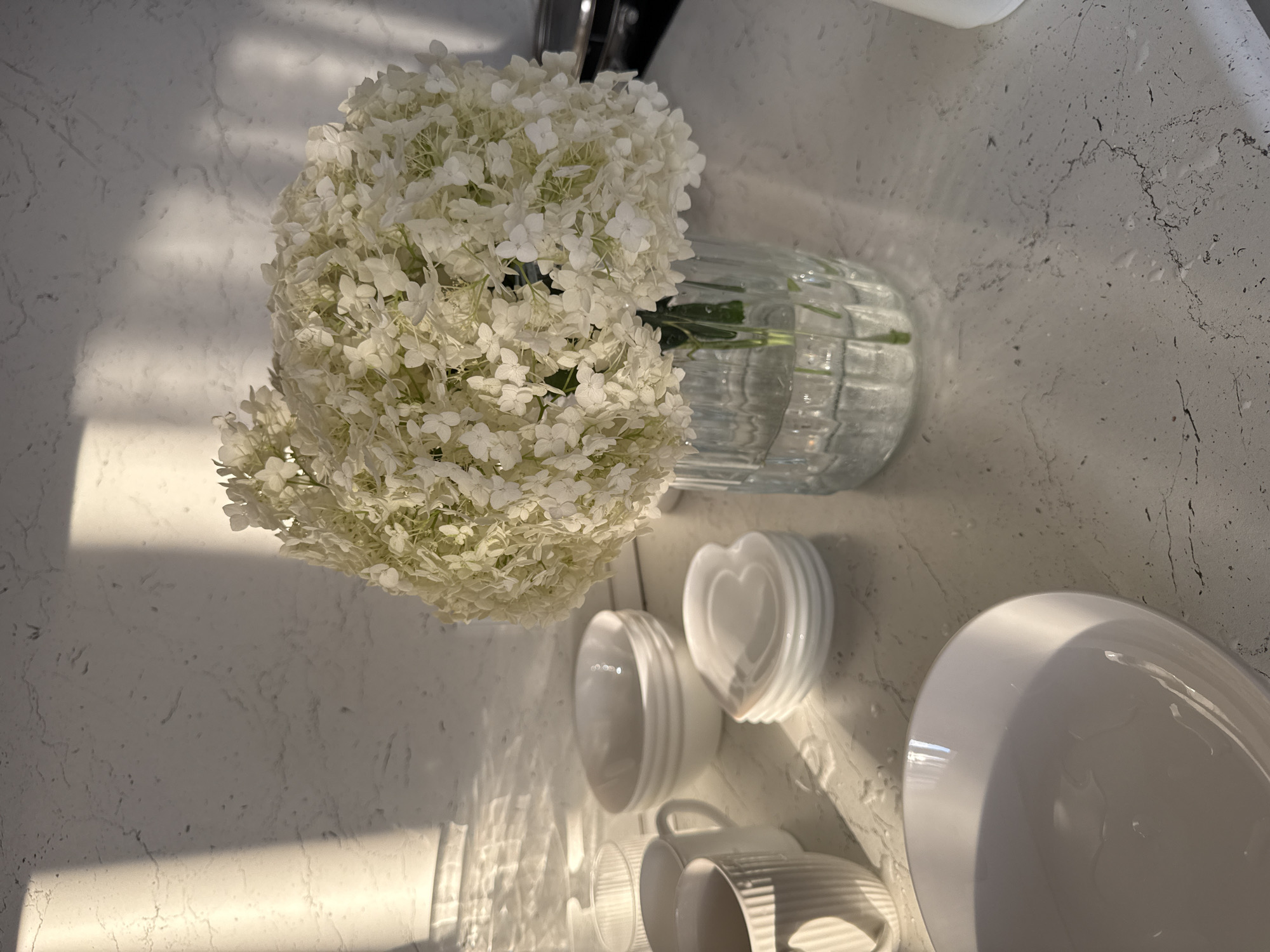 A bouquet of white hydrangeas sits in a clear glass vase on a light-colored countertop. Several white bowls and mugs are arranged around the vase. The scene is illuminated by sunlight streaming in from the left, casting shadows on the textured wall behind the countertop. The countertop itself appears to be made of a marbled material.