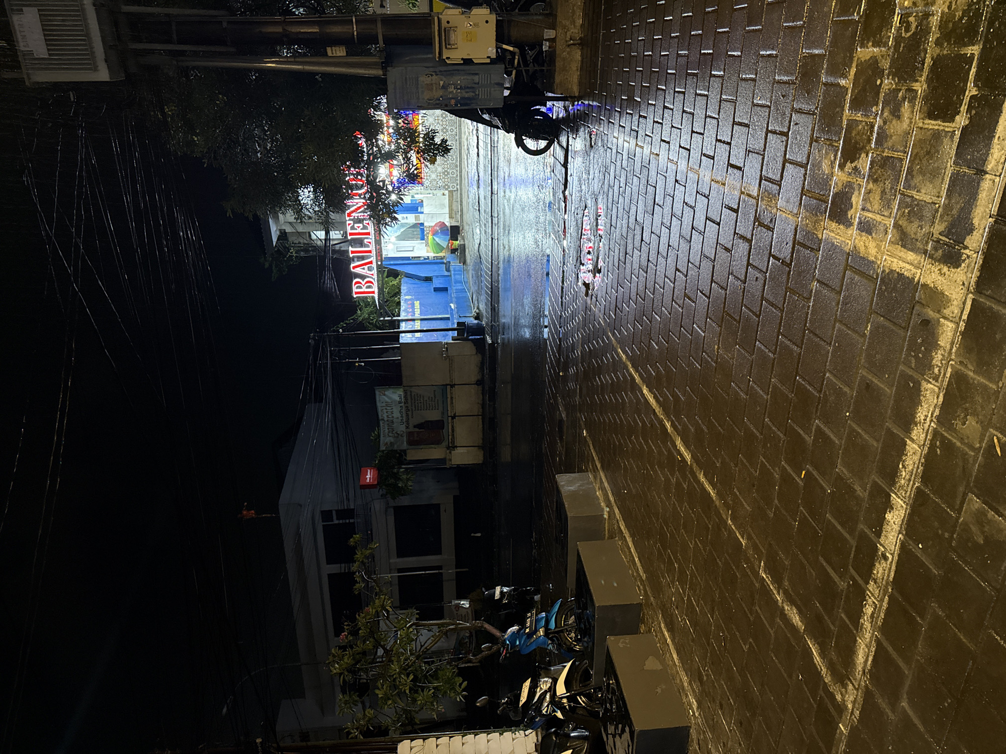 The image shows a wet, paved street in Kuta, Indonesia, at night. The street is lined with buildings, including one with a sign reading "BALENO." There are motorbikes parked along the sidewalk. The street is illuminated by streetlights and signs. There are utility poles and wires visible above the buildings.