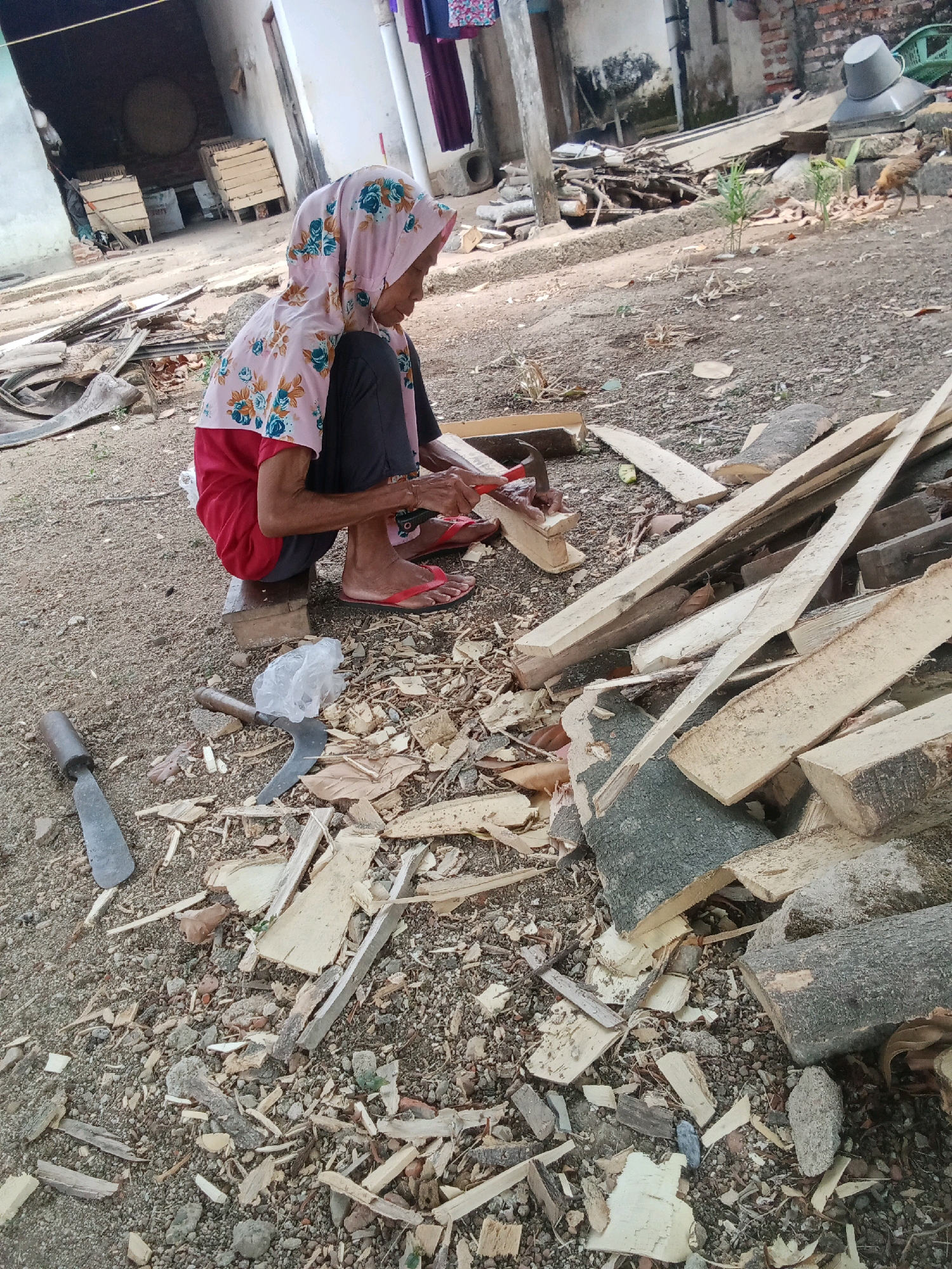An elderly Indonesian woman, wearing a pink hijab with a floral print, a pink shirt with a floral print, dark pants, and red flip-flops, is squatting on a wooden stool and carving a piece of wood with a small hand plane. She is surrounded by wood scraps and lumber in an outdoor area that appears to be a yard or workshop. In the background, there is a building with an open doorway and some wooden crates. To the right, a chicken is visible. Various tools, including a sickle-shaped knife and a chisel-like tool, are scattered on the ground. The environment is dusty and appears to be rural or semi-urban.