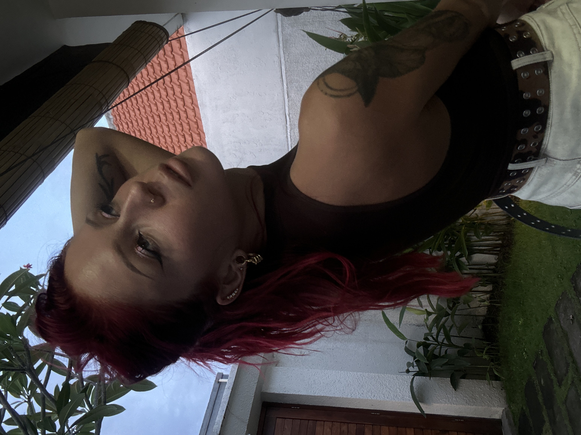 A woman with striking red hair, darker roots, and tattooed arms is the central subject of this outdoor portrait, captured from a low angle. She appears to be lying down or leaning back, looking upwards with a calm expression. Her face features a nose piercing and multiple ear piercings, including a distinctive golden ear cuff. She wears a dark, possibly brown, tank top and a patterned, studded belt over light-colored trousers or shorts.
The setting is a tropical garden or courtyard, consistent with North Kuta, Indonesia. Architectural elements in the background include a rolled-up bamboo blind, terracotta-colored roof tiles, and white stucco walls. Lush green plants and foliage are abundant, with a small plant bearing a pink flower visible near her head. A pathway of stone pavers interspersed with grass, and a partially visible wooden door, complete the scene.
The lighting is soft and even, suggesting an overcast day or late afternoon, contributing to a relaxed and tranquil atmosphere. No text is visible in the image.
