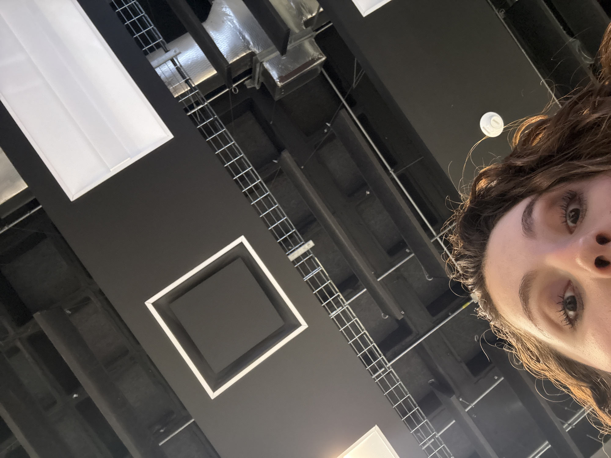 The image shows a low-angle view of a black ceiling with various features including rectangular lighting fixtures, a square recessed fixture, and visible overhead utilities like conduit or cable trays. A woman's face is prominently visible in the right side of the frame.
The location is indeterminate from the image. The city and country cannot be determined from the given information.