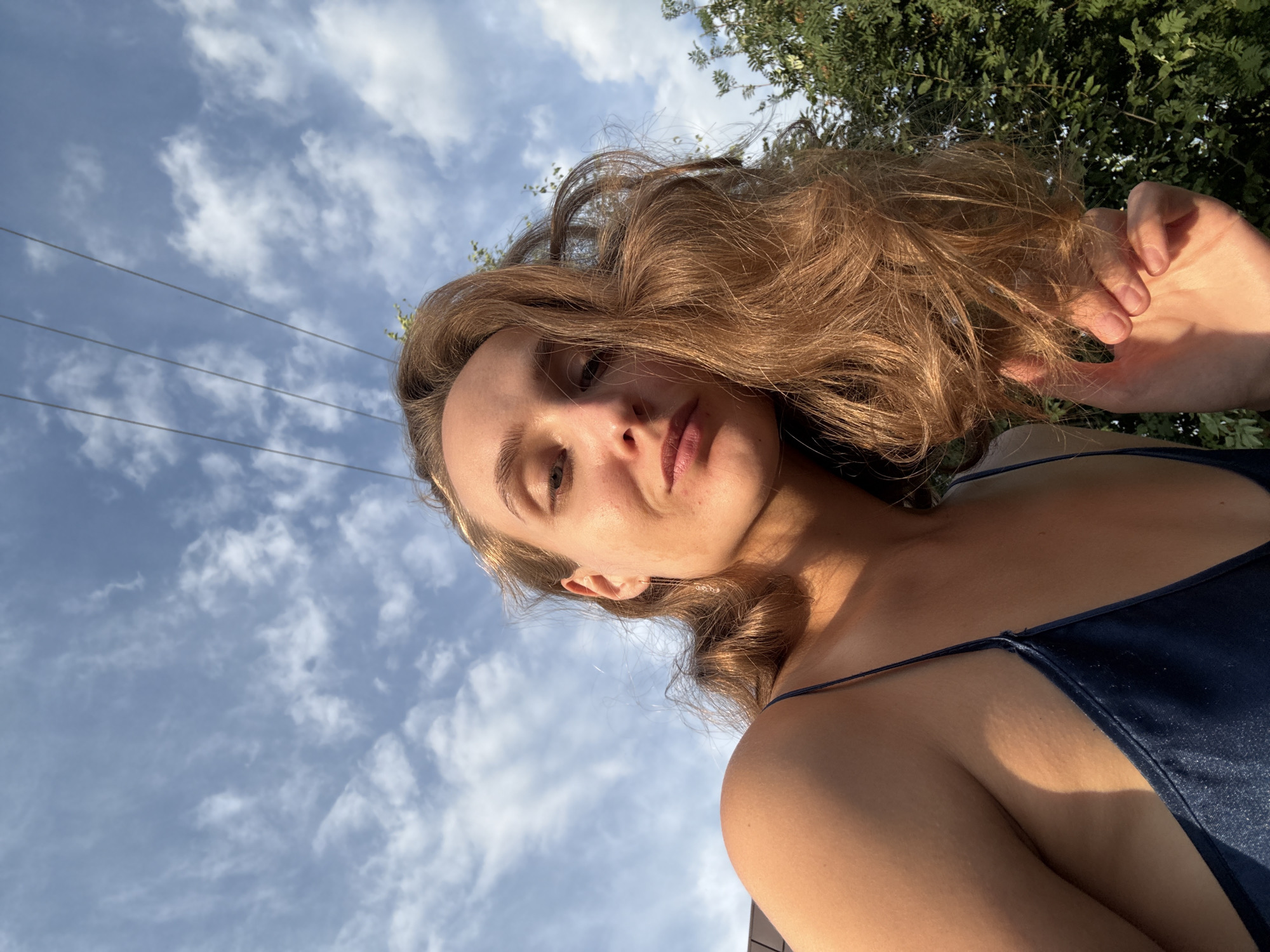A young woman with long, wavy brown hair is pictured from a low angle, looking up at the camera. She is wearing a dark blue spaghetti strap dress. The background consists of a blue sky with scattered white clouds and several power lines running horizontally across the upper portion of the frame. To the right, green foliage from trees is visible. The lighting suggests it is late afternoon or early evening, with the sun casting a warm glow on the woman's skin. Her facial expression is neutral, with her lips slightly pursed. The scene is outdoors, and there is no indication of the specific location within Sharkan, Russia.