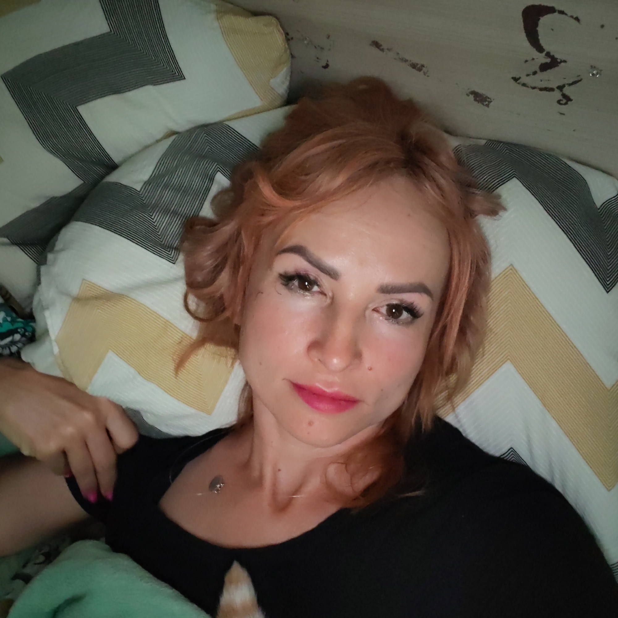 A single adult female is depicted lying supine in an indoor setting, identified as the city of Chaikovsky, Russia. Her head is positioned on a pillow with a white cover featuring grey and light yellow chevron patterns. She has light reddish-brown hair, styled loosely, and is looking directly into the camera. Her facial features include dark eyes, pronounced eyebrows, and bright pink lipstick. She wears a black short-sleeved top and a silver-toned pendant necklace. Her left hand, adorned with pink nail polish, is visible on her left side near her chest. A second pillow with grey chevron patterns is partially visible above and to her left. A light teal blanket is present in the lower left portion of the frame. The upper right background displays a light-colored surface with several irregular dark marks.