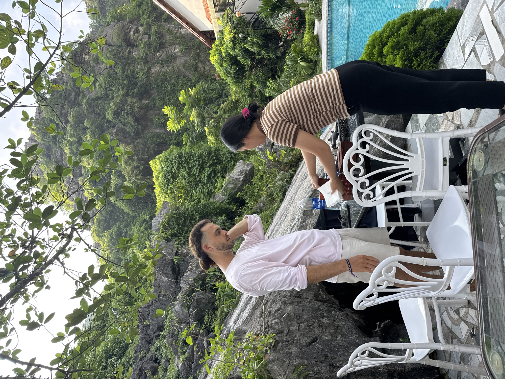 In a tranquil outdoor setting, likely within Nam Hoa Lư Ward, Vietnam, a man with a man bun, wearing a pale pink long-sleeved shirt and white shorts, stands with his hand on his neck, appearing contemplative. He is positioned near a rocky outcrop adorned with lush greenery. To his right, a woman in a brown and white striped shirt and black pants is actively engaged with items on a table, possibly preparing food or drinks. The scene is further enhanced by a sparkling blue swimming pool in the background, a villa with a tiled roof, and elegant white chairs arranged around tables. The surrounding environment is dominated by the majestic presence of verdant, rocky hills, creating a serene and picturesque atmosphere.