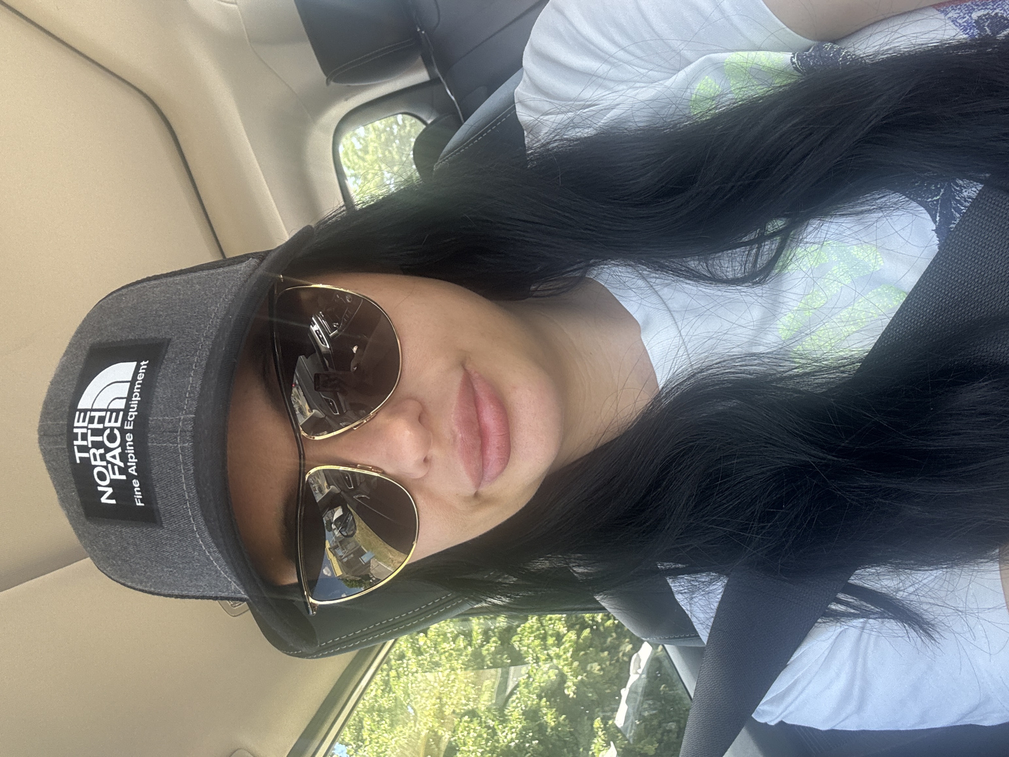 The image is a selfie taken inside a vehicle. A woman with long, black hair and sunglasses is wearing a gray North Face cap and a white t-shirt. She is also wearing a seatbelt. Through the car window, green trees are visible. The location is likely an urban area, but the specific city is unclear.