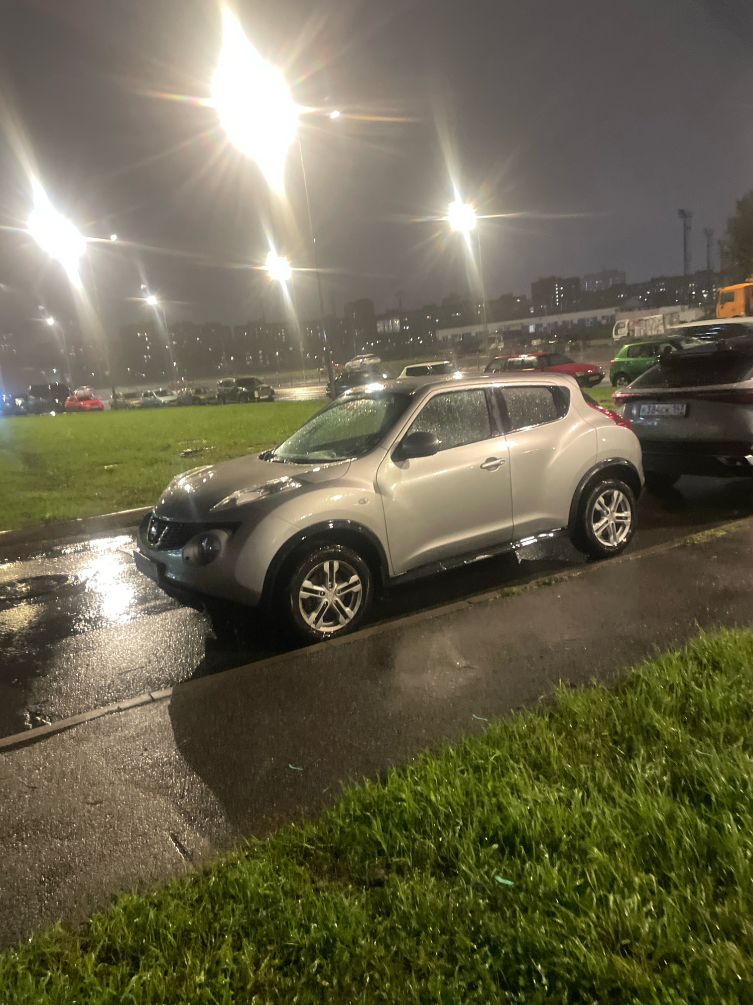 The image depicts a street scene at night in an urban environment, likely a parking lot or roadside, with wet asphalt suggesting recent rain. The foreground is dominated by the front quarter of a silver Nissan Juke, parked on the right side of the frame. The car has water droplets on its surface. To the right of the Nissan Juke, a portion of a darker colored car is visible. In the background, multiple other vehicles are parked in rows on a grassy area. Streetlights cast a bright, diffused glow, creating lens flare effects in the image. The sky is dark and overcast. The location is identified as Unknown, Russia.