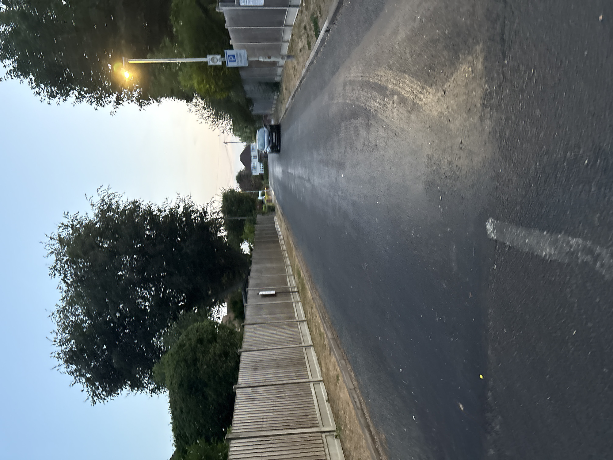 A black car is parked on the side of a street in Chelmsford, United Kingdom. The street is paved with asphalt and has a curb on the right side. A wooden fence runs along the left side of the street. Trees and houses are visible in the background. The sky is clear and blue.