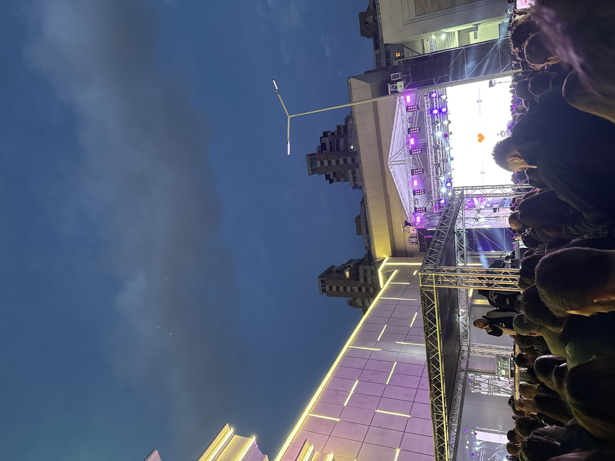 The image captures an outdoor event at night in Yerevan, Armenia. A crowd of people are gathered in front of a stage, which is illuminated with bright lights, including purple ones. There's a large screen on the stage displaying the logo of "Coffee House". The architecture of the surrounding buildings is visible, including a tall, modern building lit with yellow and purple accents, and another multi-story building further in the background. The sky is a deep blue, with some clouds visible. A Y-shaped light pole is also visible. The crowd is dense and the people are facing towards the stage, suggesting they are watching an event or performance.