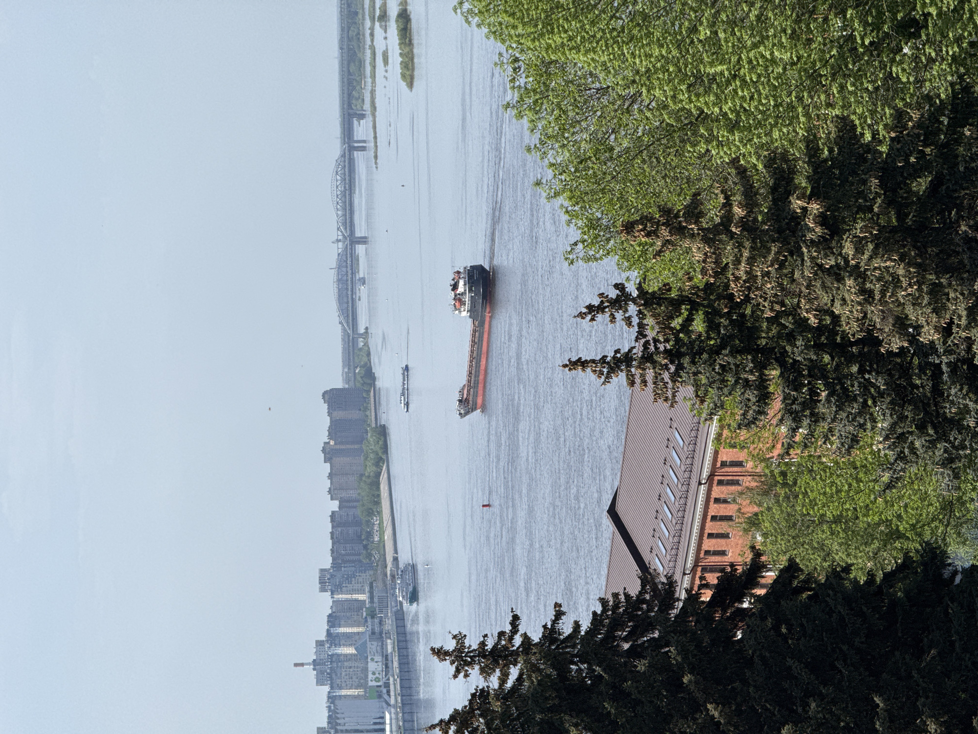 Here's a description of the image:
The image shows a wide river with a large cargo ship moving along it. A second, smaller boat is also visible on the water. A city skyline runs along the left side of the riverbank, featuring buildings of various sizes and architectural styles. Two large bridges span the river in the distance. The foreground is dominated by trees and greenery, with a building visible nestled among them. The building is constructed of red brick, with a brown roof and several windows visible.
It appears to be the Volga River in Nizhny Novgorod, Russia, due to the types of bridges and buildings shown, as well as the presence of large cargo ships on the river.