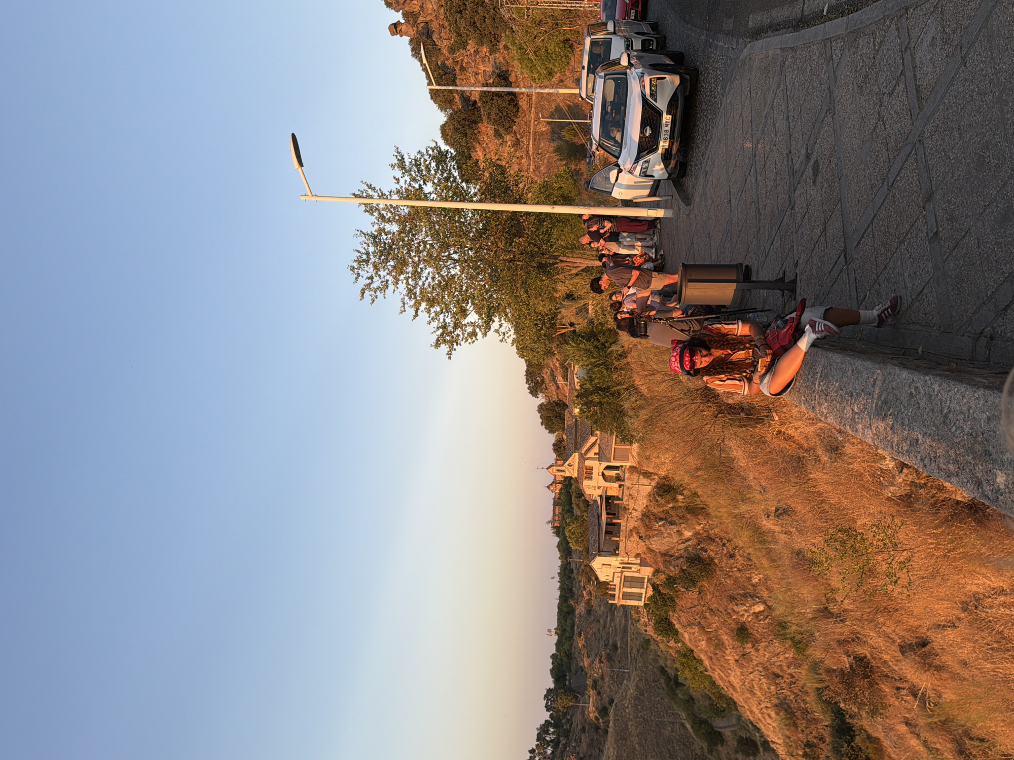The image depicts a group of people gathered along a stone wall overlooking a landscape in Toledo, Spain, likely near the Mirador del Valle viewpoint. The setting is illuminated by the warm, golden light of late afternoon or early evening. A young woman in a pink hat and striped shirt is seated on the wall in the foreground, looking at her phone. Behind her, a line of cars is parked on the right side of a paved walkway. In the background, traditional Spanish architecture, including buildings with red tile roofs and a church with a steeple, is visible perched on a hillside. The surrounding terrain is dry and covered in sparse vegetation, characteristic of the Castilian landscape. The sky is a clear, pale blue, transitioning to a warmer hue near the horizon.