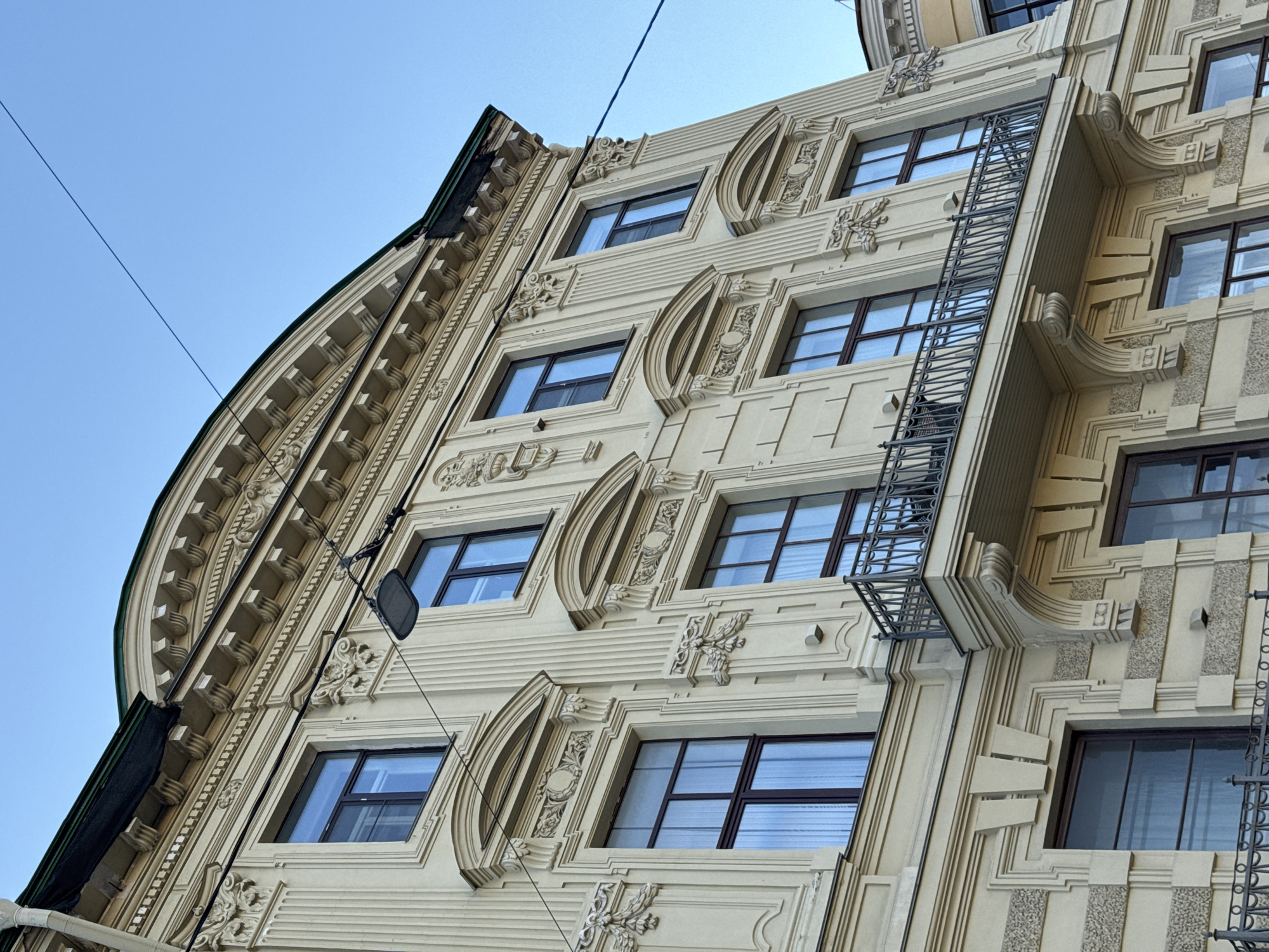 An upward-angled shot captures the ornate facade of a multi-story building against a clear, bright blue sky in Saint Petersburg, Russia. The building, rendered in a light, creamy yellow or beige, exhibits intricate architectural details typical of historic European styles, possibly Art Deco or Neoclassical.
The leftmost section of the building features a grand, curved or turret-like structure with a green roofline and repetitive decorative moldings. The main facade is adorned with a rich array of embellishments, including pilasters, horizontal bands, elaborate sculptural reliefs resembling floral motifs or cartouches, and geometric patterns. Many windows with dark frames and reflective glass are visible, showcasing faint reflections of the sky or interior elements.
A prominent feature is an ornate metal fire escape or ladder running vertically up the right-center portion of the building, adding a functional yet decorative element to the structure. Some sections of the facade display textured finishes, possibly pebble dash or roughcast. Thin black wires crisscross parts of the sky and the building, with a small black object attached to one wire in the mid-left. The scene is bathed in bright daylight, indicated by the clear shadows and uniformly blue sky, suggesting a sunny day. No people or visible text are present.