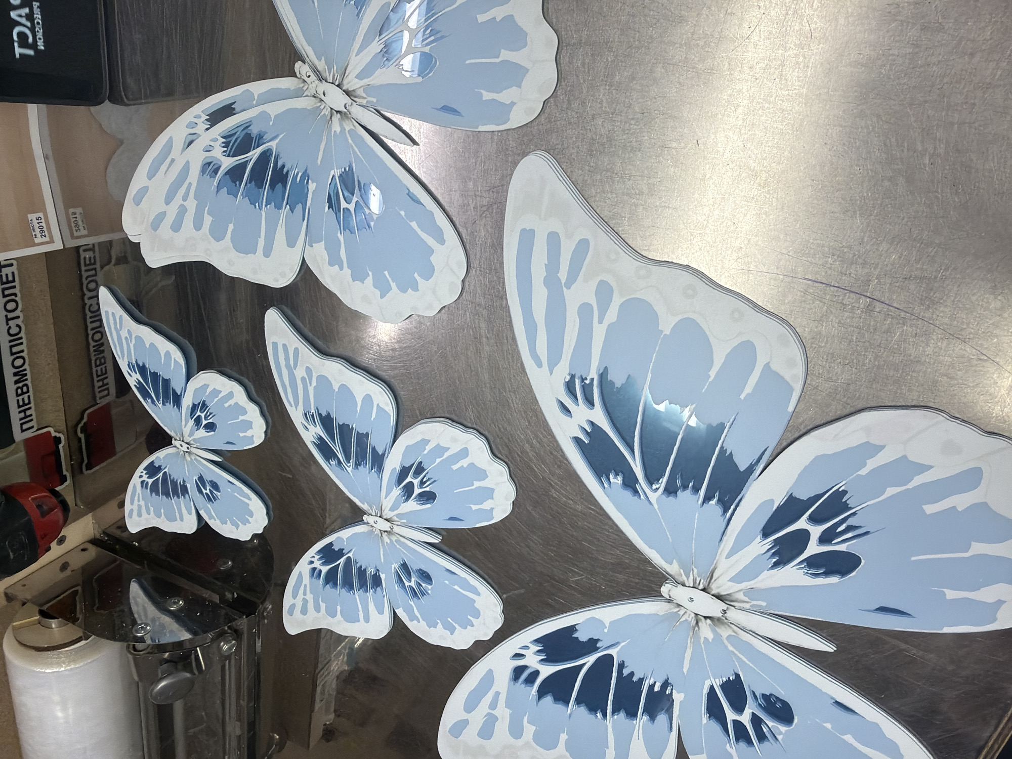 The image displays multiple stylized butterfly cutouts resting on a brushed metallic surface, likely in an industrial or workshop setting in Ternopil, Ukraine. There are at least five butterflies of varying sizes, predominantly light blue and white with darker blue patterns, featuring a reflective, layered appearance that suggests a three-dimensional design. Their glossy surfaces reflect ambient light and hints of the surroundings.
The metallic background exhibits numerous parallel scratches and scuffs, indicating wear. A faint purple line is visible on this surface in the upper right. In the bottom-left corner, a spool of white material, possibly plastic film, is visible alongside a section of metallic machinery. Above this, a red and black object, likely part of a tool, is partially seen. A black box or container is also visible in the top-left, reflecting part of its label.
No human subjects are present, and no specific activity is taking place, suggesting the butterflies are decorative items, possibly awaiting assembly or display.
Visible text includes:
- "29015" on a brown label/sticker.
- "NPOCTA" (likely "ПРОСТА", meaning "SIMPLE") on a white label.
- Reversed text "ПНЕВМОПИСТОЛЕТ" (PNEUMATIC PISTOL/AIR GUN) on a vertical object.
- Reversed text "ТОЛСТ" (possibly "ТОЛСТЫЙ," meaning "THICK") on a black object.