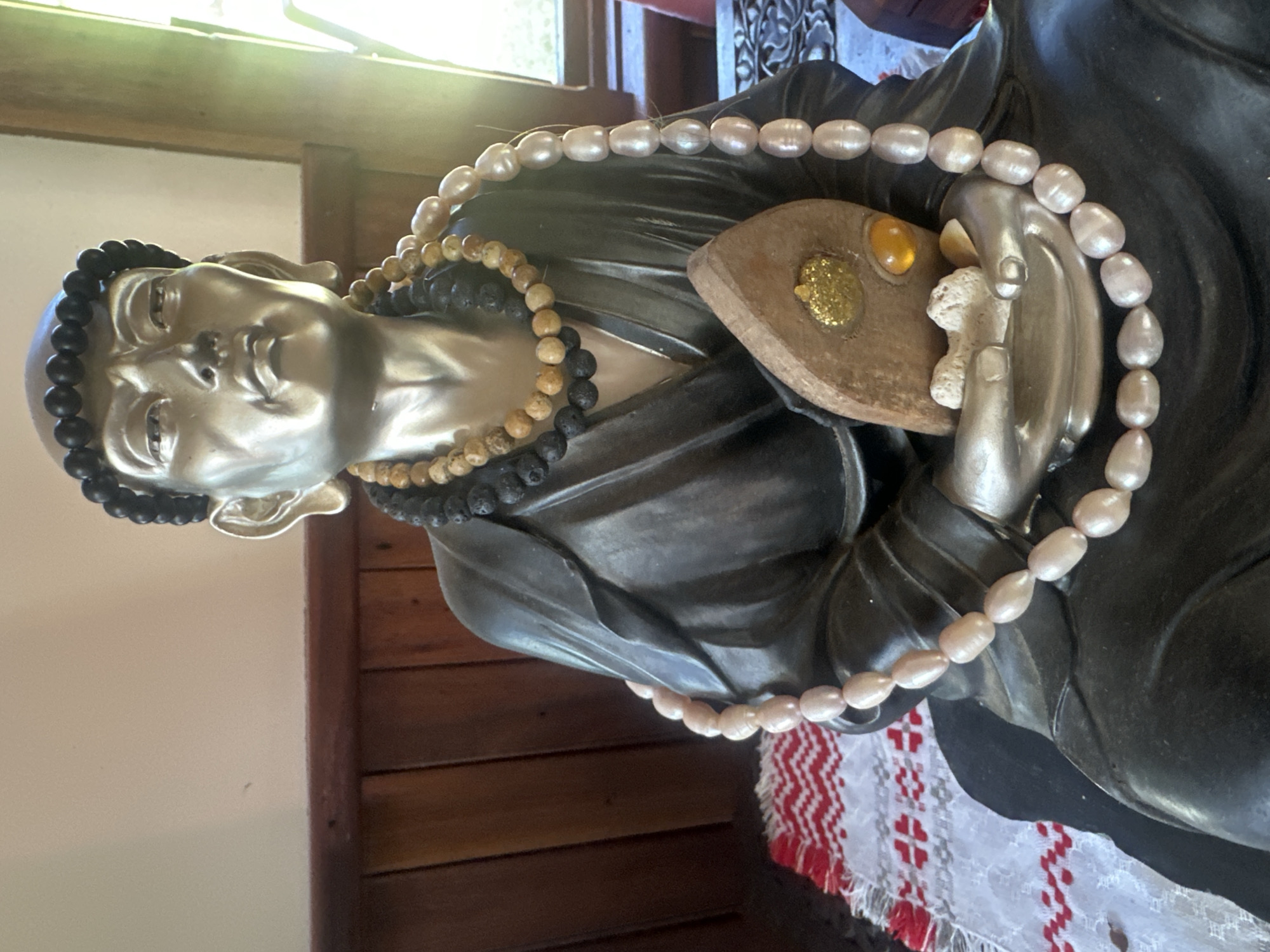 The image displays a close-up view of a statue of a seated figure, likely a monk or spiritual leader, adorned with multiple necklaces. The statue is made of a metallic material, with a silver-toned head and face, and a dark, draped robe. The figure wears a circular string of black beads around its head and two layered necklaces around its neck. The top necklace consists of dark, porous beads, possibly lava beads, interspersed with lighter, stone-like beads. The lower necklace is a string of large, oval, pale pink pearls. The statue's hands are clasped together in its lap, holding a small, triangular, brown object that appears to be a piece of wood or stone. This object has a small, circular, gold-colored embellishment with glitter and a small, amber-colored gem. The statue is seated on a dark base, which rests on a white cloth with a red and white geometric pattern. The background consists of dark wooden paneling and a light-colored wall. A window, possibly to the right of the statue, allows sunlight to illuminate the scene. There are no people present in the image. The image does not provide any information that would allow for the determination of its location within the city of Thora, Australia.