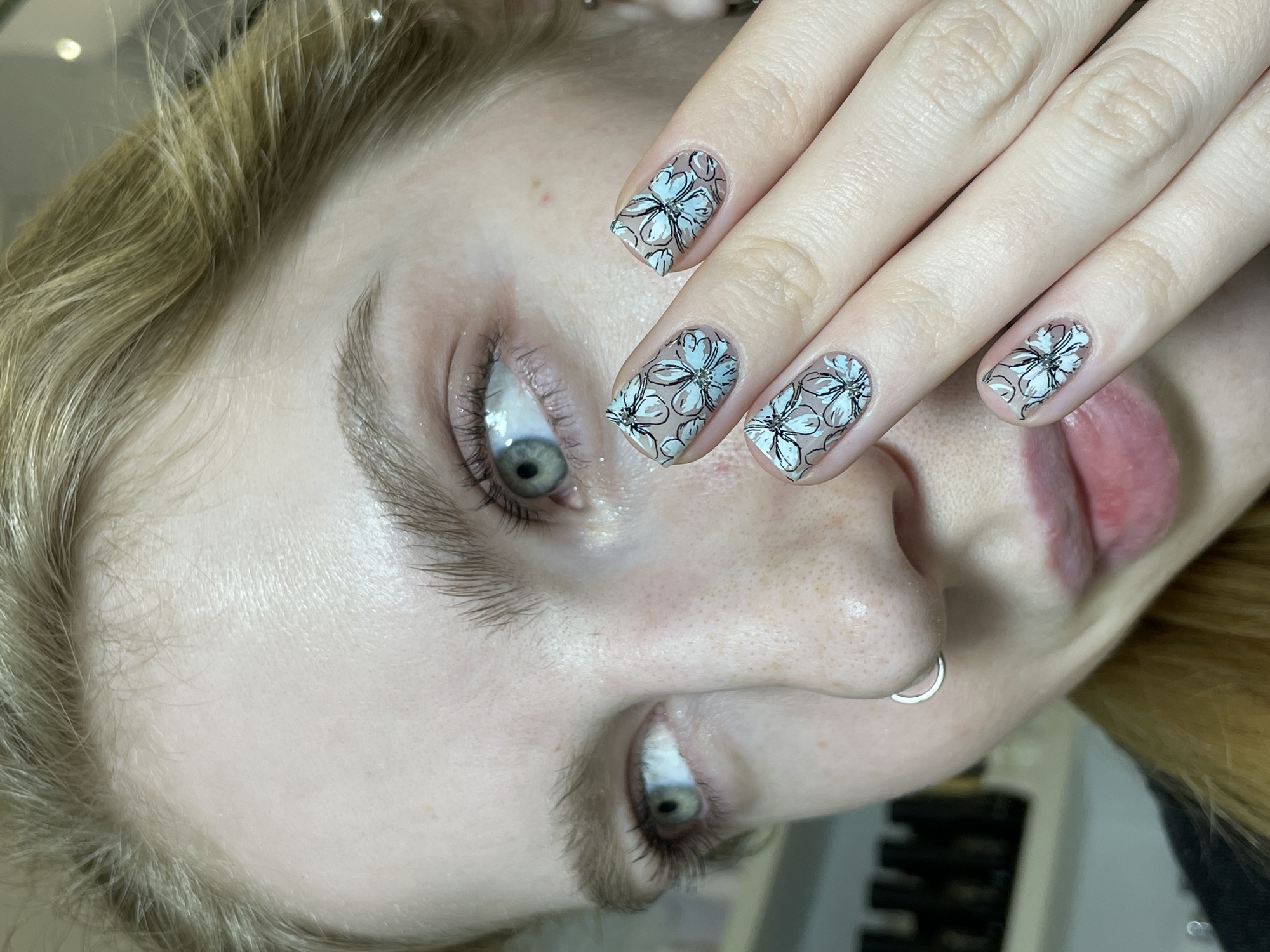 A close-up, rotated view shows a light-skinned person with blonde hair and light-colored eyes. The person has a silver hoop nose ring. Their fingernails are painted with a nude base color and floral designs. The design appears to be white flowers with black outlines and small gemstones in the center. The background is indistinct. No information suggests this image was taken in Izhevsk, Russia.