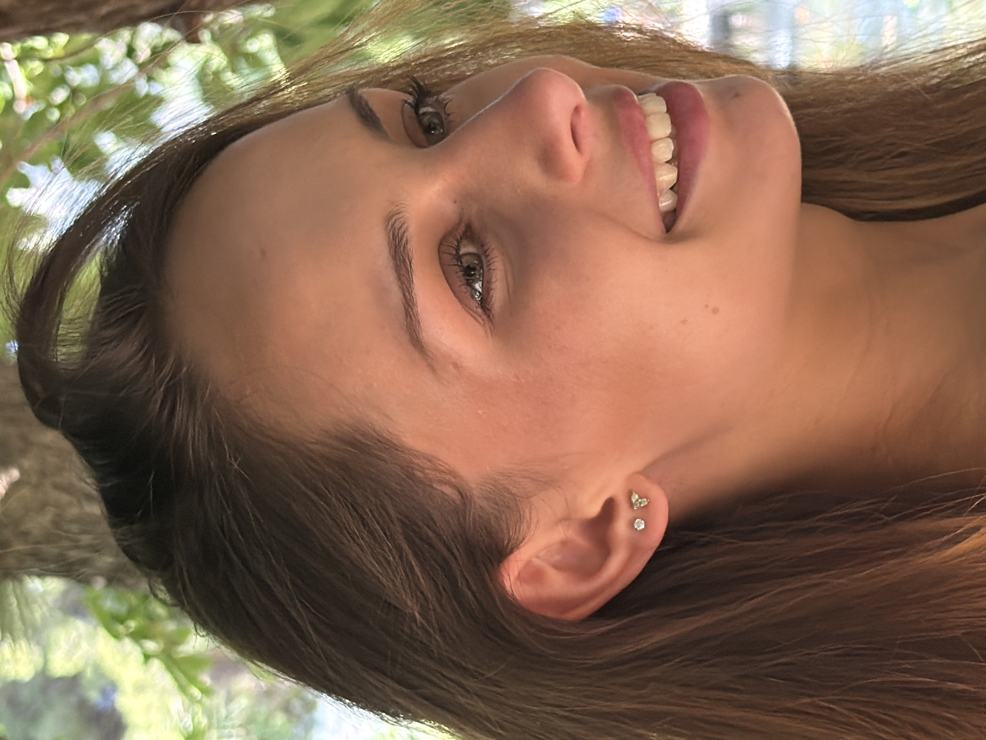 A young woman with light brown hair is smiling and looking upward. She has fair skin, light-colored eyes, and is wearing earrings in her left ear. The background features foliage, suggesting an outdoor setting. There is no clear indication of the location being Bunyola, Spain, from the provided image.