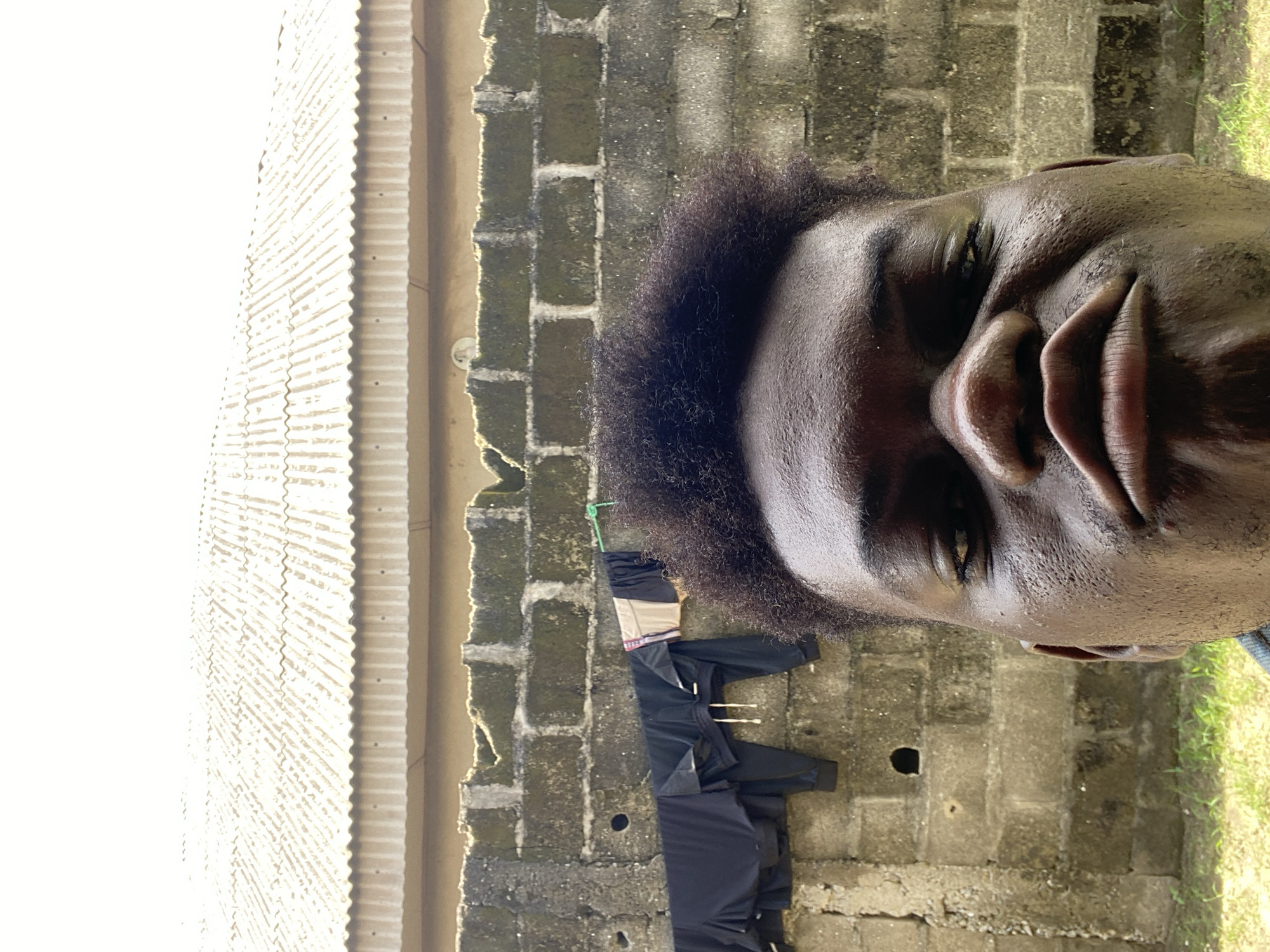 A close-up portrait of a dark-skinned man with an afro is positioned in front of a weathered brick wall and a corrugated metal roof. Laundry hangs on a clothesline attached to the wall. The location is the city of Unknown, Unknown.