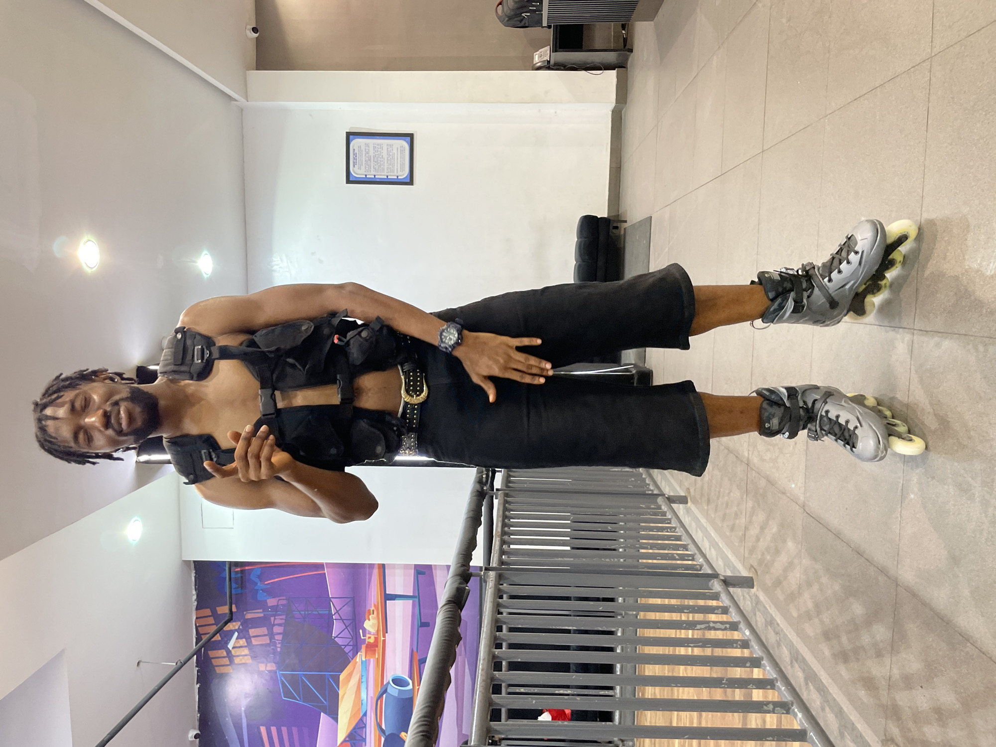 A Black male, appearing to be in his late 20s to early 30s, stands facing the camera in what appears to be an indoor roller skating rink. He is wearing a black tactical vest over a bare torso, black shorts that fall below the knee, a black belt with a gold buckle, and silver roller skates. His hair is styled in dreadlocks. He is pointing his right index finger towards the camera with his left hand resting on his left thigh. The background features a mural on the left wall depicting a cityscape with a ramp and a table with food, and a white wall with a framed document on the right. A metal railing is visible in the foreground. The flooring is tiled. The scene is well-lit, suggesting an indoor public space. The location in the city of Moba, Nigeria, is not ascertainable from the provided image alone.