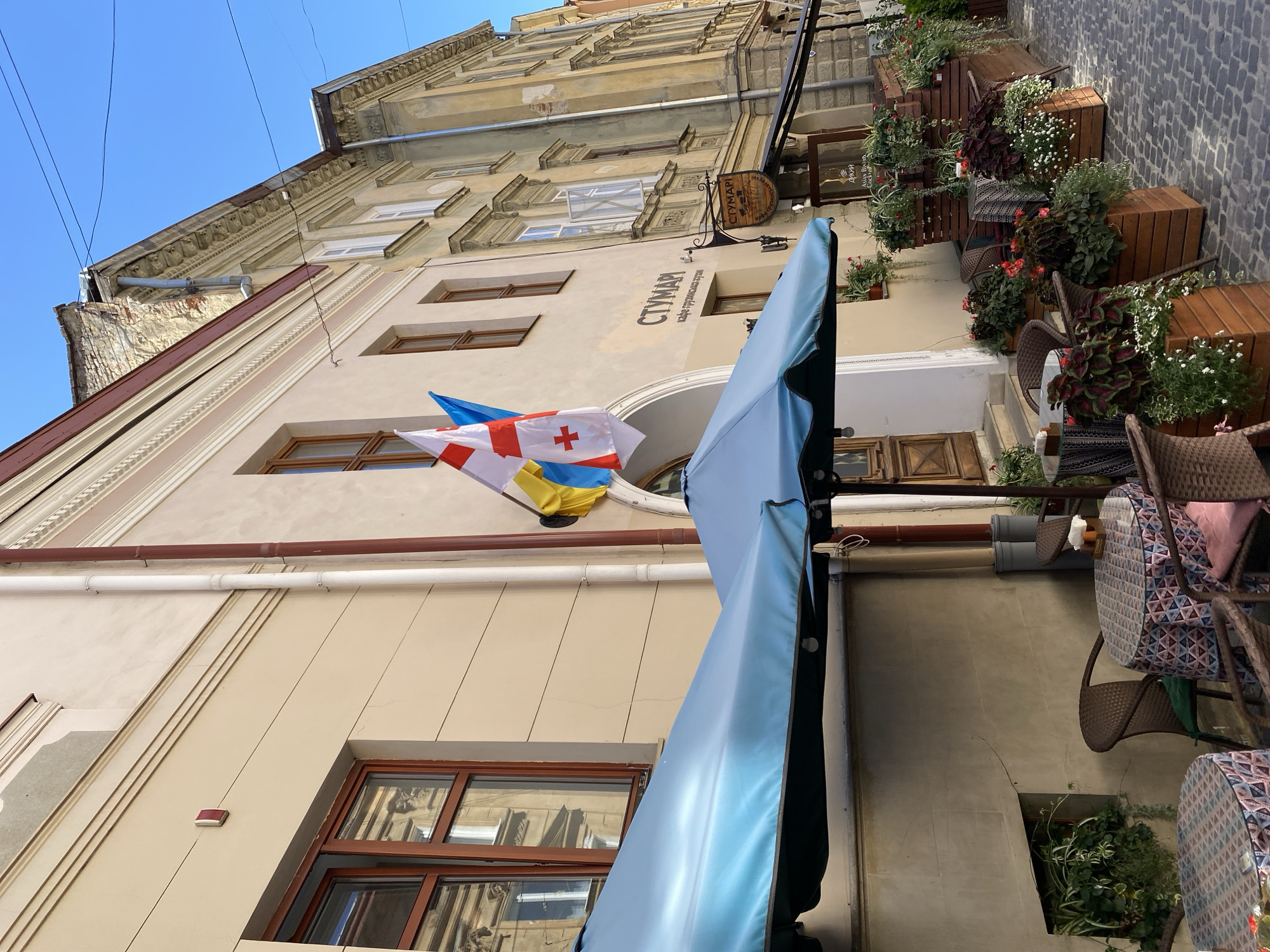 In Lviv, Ukraine, on a sunlit day, an outdoor seating area of a cafe is set up on a cobblestone street. Two blue umbrellas provide shade over tables adorned with colorful, patterned tablecloths. Wicker chairs are arranged around the tables. Planters filled with lush greenery and red and white flowers line the sidewalk, creating a vibrant border. The cafe building, with its light-colored facade, features several windows and architectural details. Two flags, one of Georgia and one of Ukraine, are displayed prominently on the building's exterior. A sign for the cafe, "СТУМАРІ", is visible, along with smaller signage indicating "Asian Bistro Cocktails". The scene captures a tranquil moment in a European city, with the cafe and its inviting outdoor space bathed in natural light.